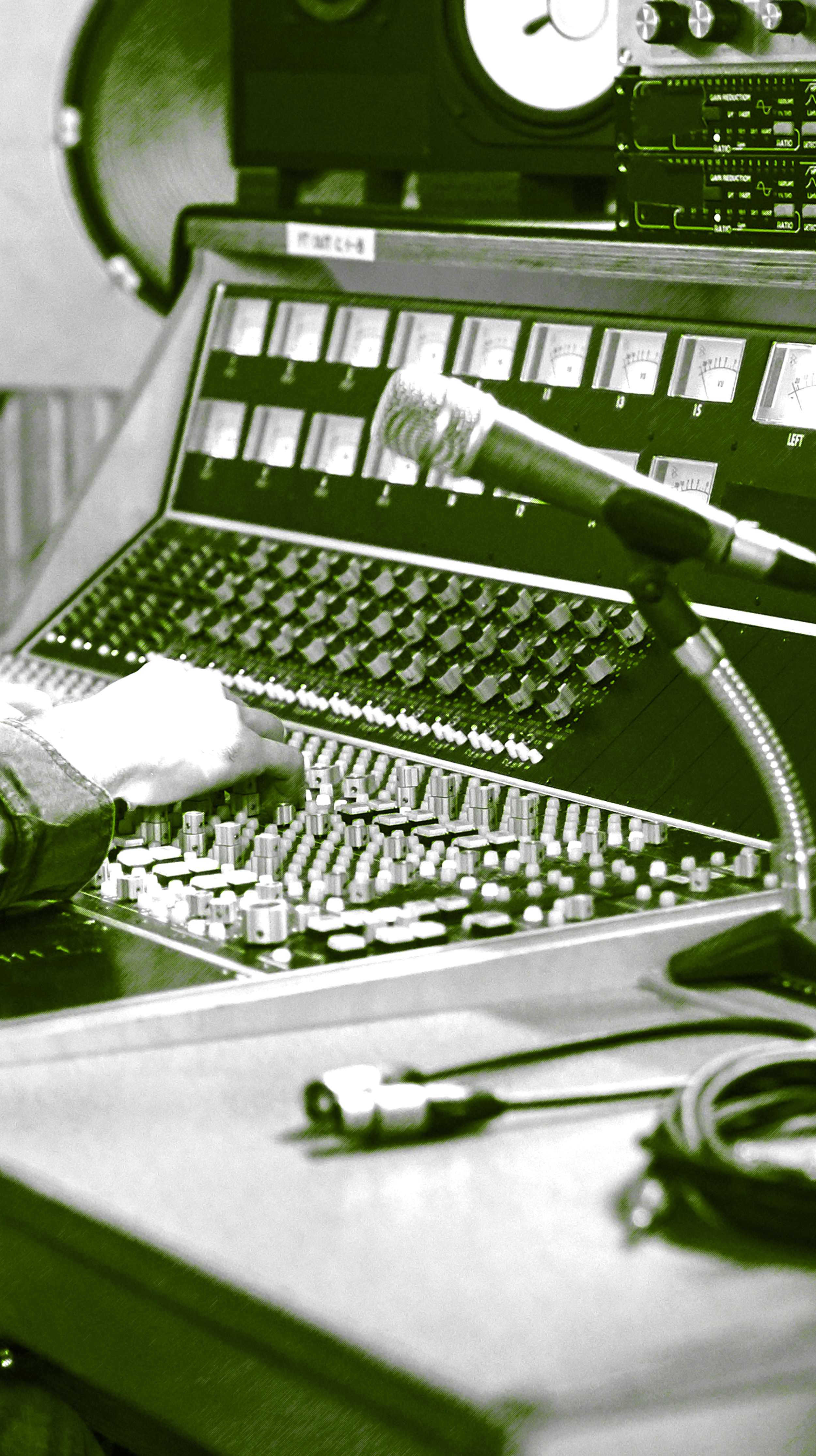 Mesa de controle com equipamento de áudio, mixer com vários botões, controles e faders, além de um microfone ligado a um cabo, sendo operada por uma pessoa. Há também caixas de som ao fundo.