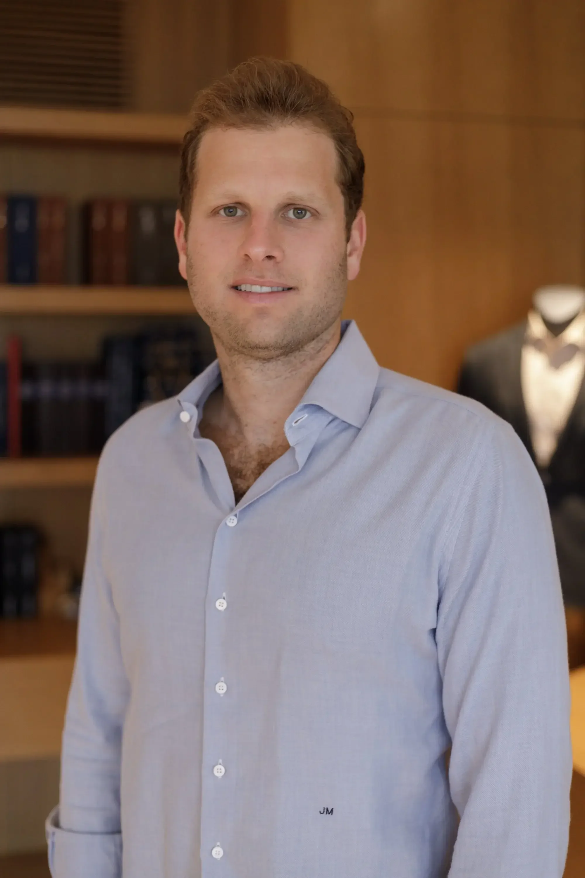A portrait of a young man with short, light brown hair, wearing a light blue dress shirt, standing in an elegant room with wooden shelves and a statue of a tuxedoed figure in the background.