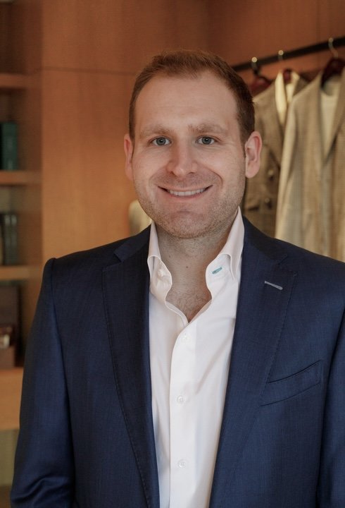 A smiling man in a dark suit and white shirt standing indoors with wooden shelves and hanging clothing behind him.