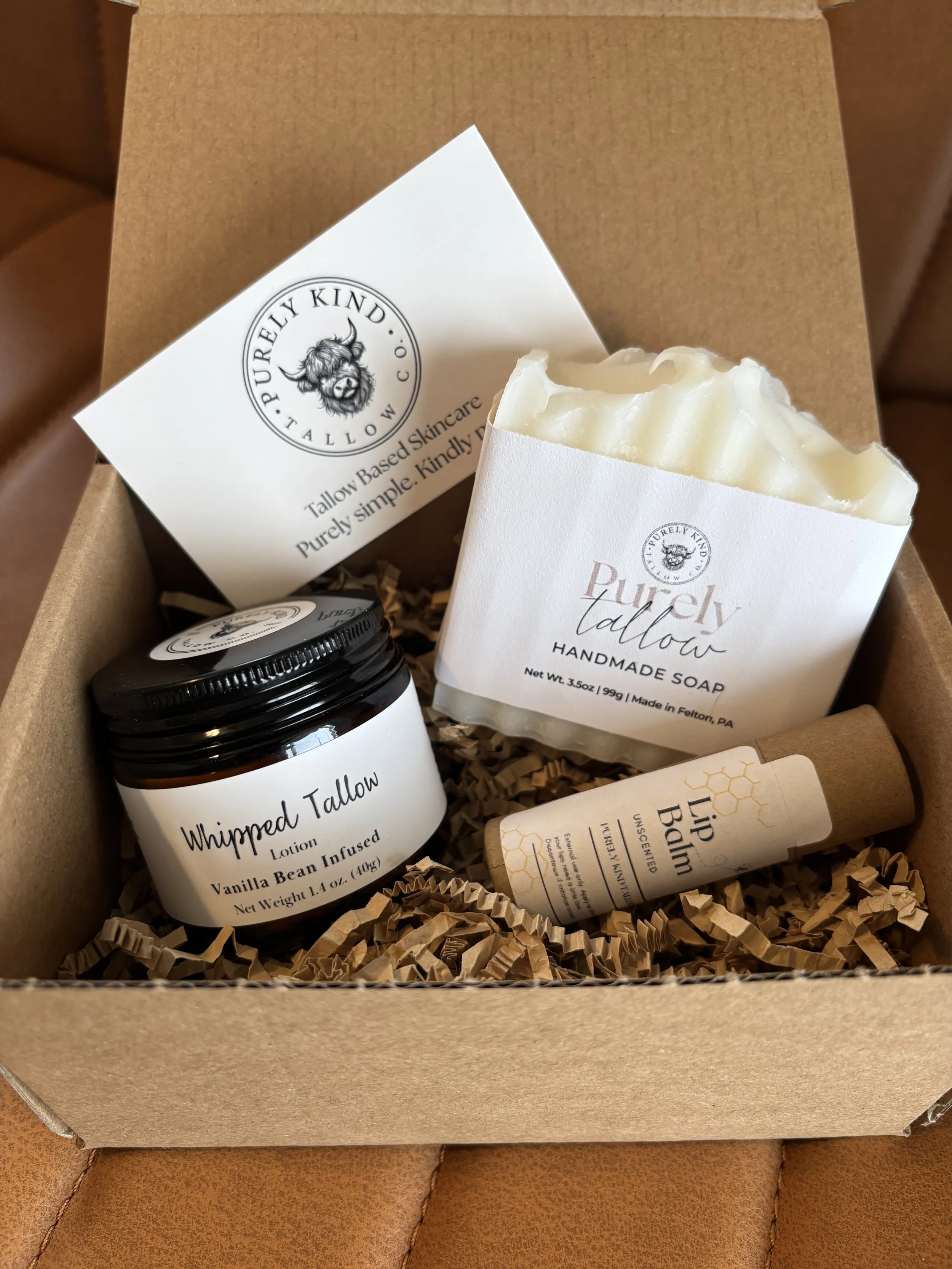 A gift box containing handmade soap, vanilla bean lotion, lip balm, and a card from Purely Kynd, with shredded paper filler.
