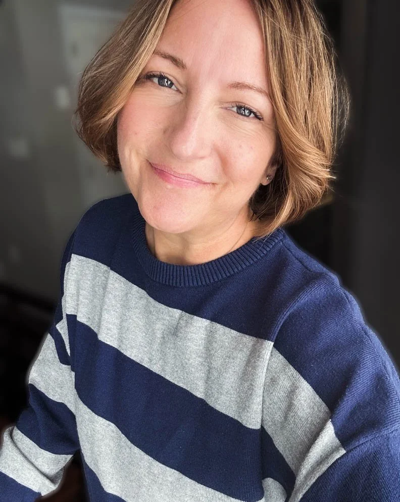 A woman with short, wavy, light brown hair smiling and wearing a navy and gray striped sweater.