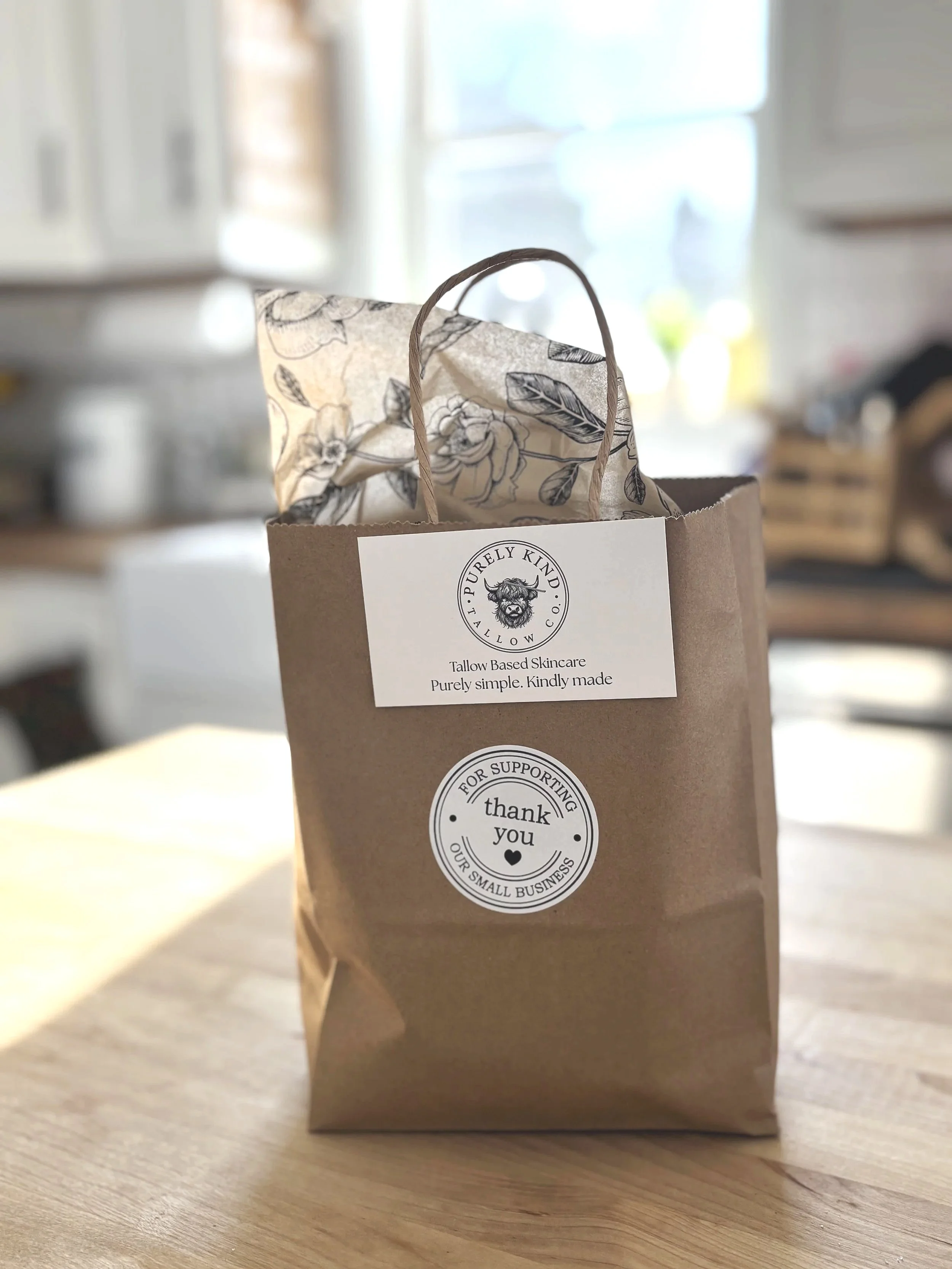 A brown paper gift bag on a wooden table with black and white stickers that read 'Purely Kind Tellow Co' and 'Thank You for Supporting Our Small Business'. Inside the bag, there is tissue paper with a floral design.