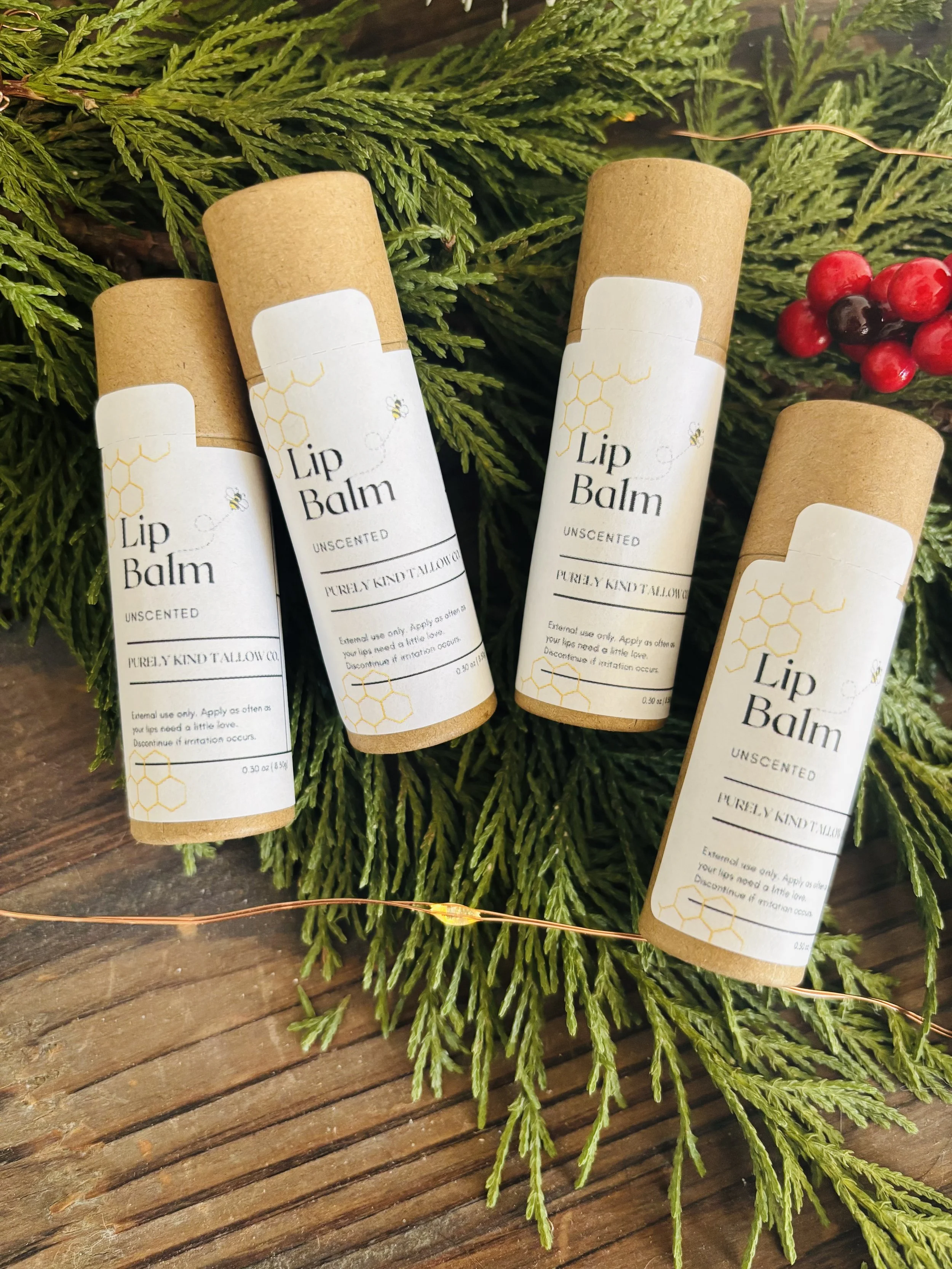 Four tubes of unscented lip balm placed on green pine branches and a wooden surface, with a small cluster of red berries in the background.