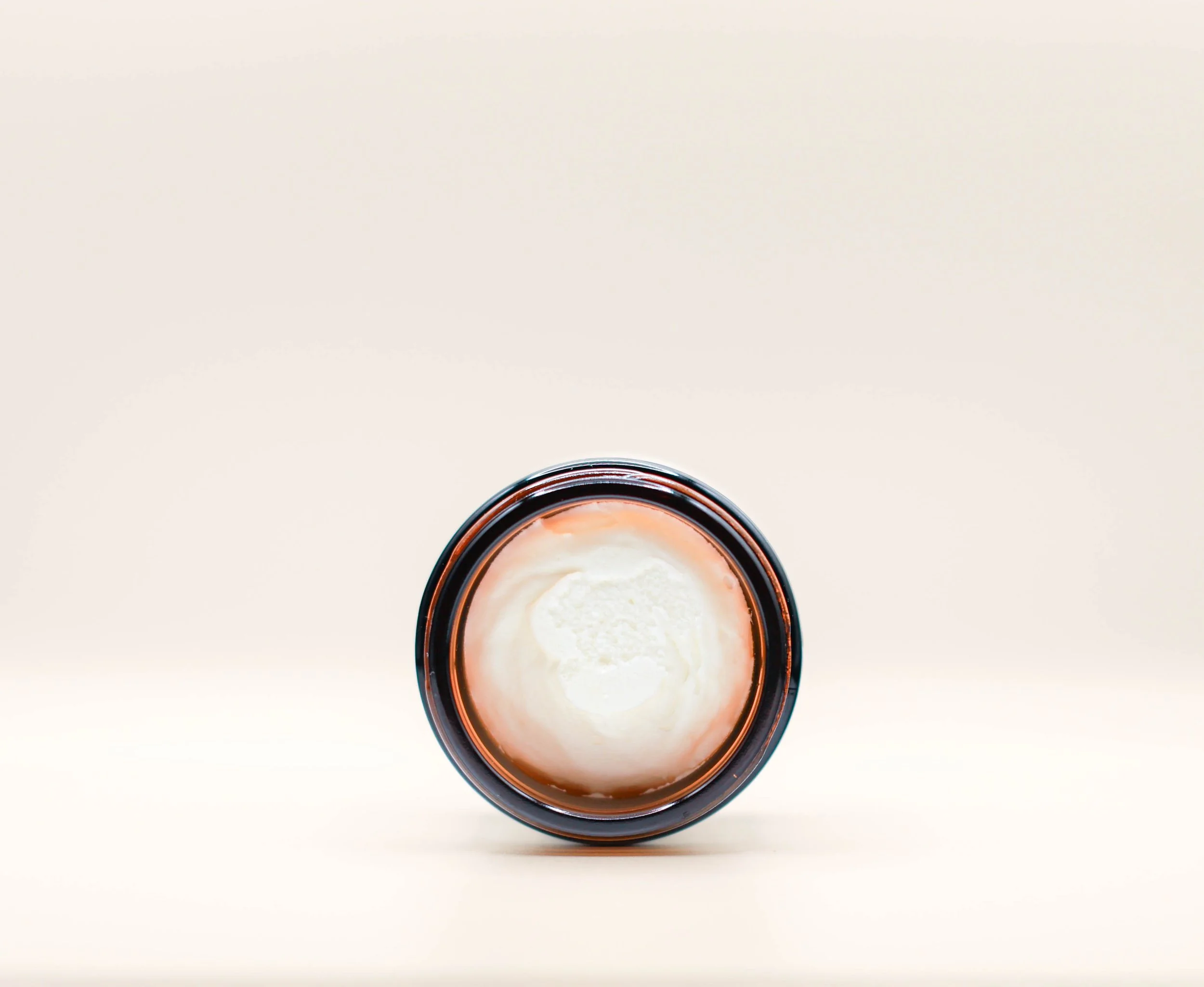 Top view of an open jar of creamy white substance, possibly lotion or moisturizer, on a plain light-colored background.