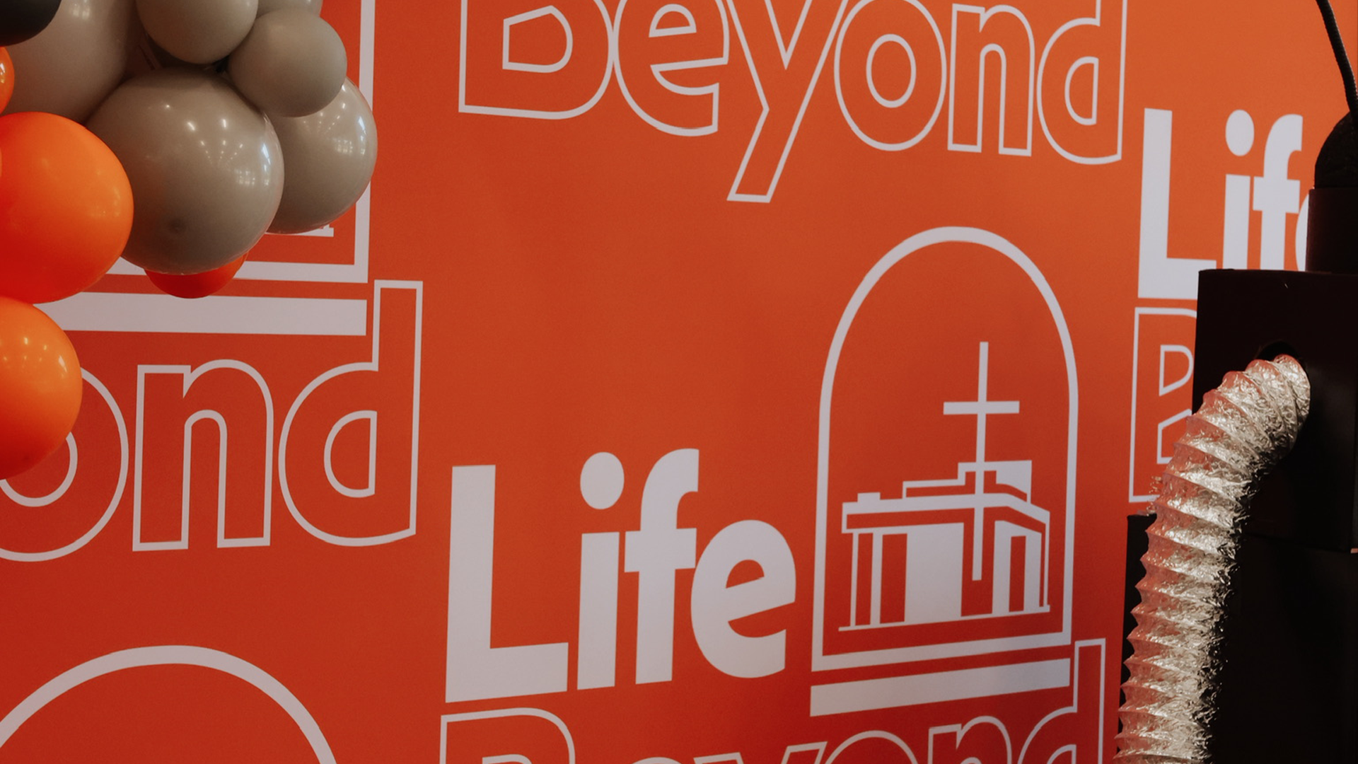 Close-up of an orange background with white text and a graphic that reads 'Beyond Life' and features an illustration of a gift box with a bow, partially visible on the right. Some balloons in beige and orange are seen in the upper left corner, and a metallic, flexible tube is on the right side.