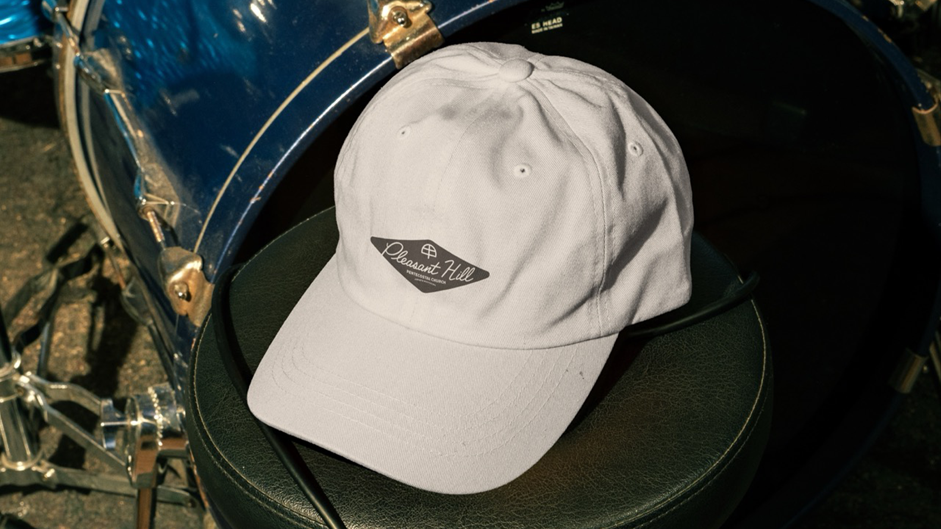 A white baseball cap with a black and white embroidered patch that reads "Pleasant Hill Baptist Church" resting on a black seat. The seat is attached to a vintage blue bicycle with a black frame and chrome details.