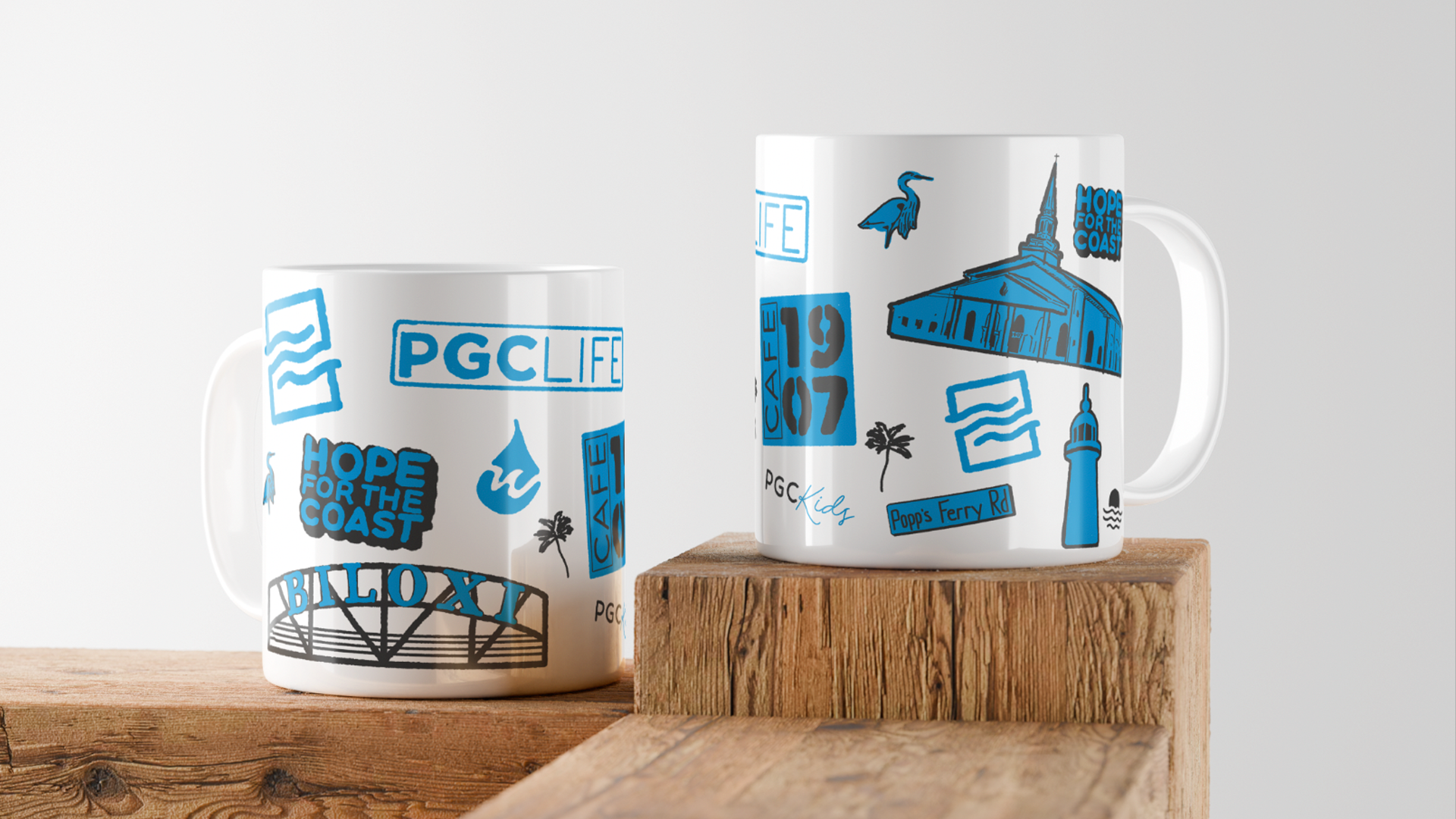 White ceramic mugs with blue and black illustrations and text featuring landmarks, coastal theme, and street names, placed on wooden blocks.