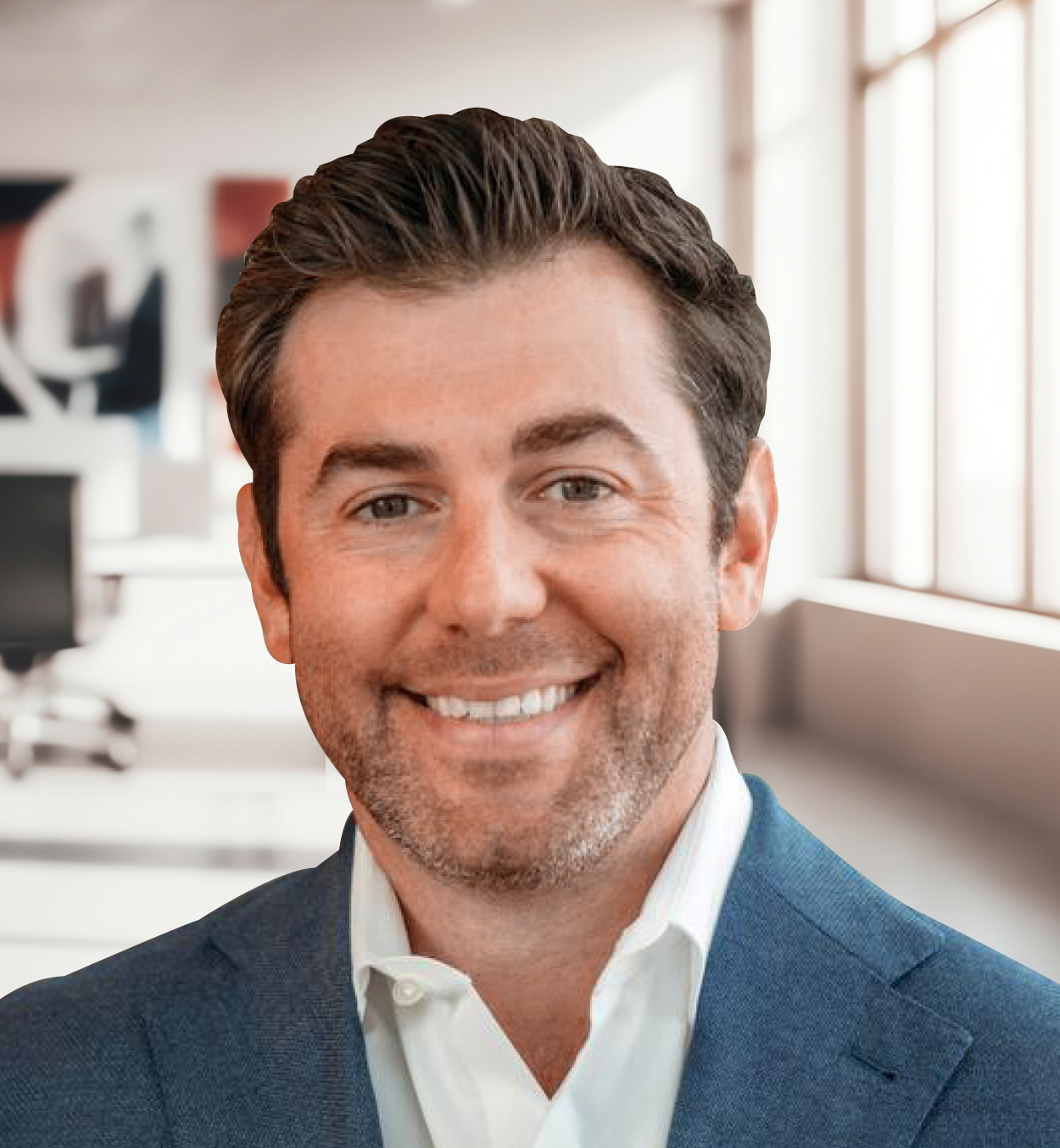 A man in a suit smiling in an office setting with large windows in the background.