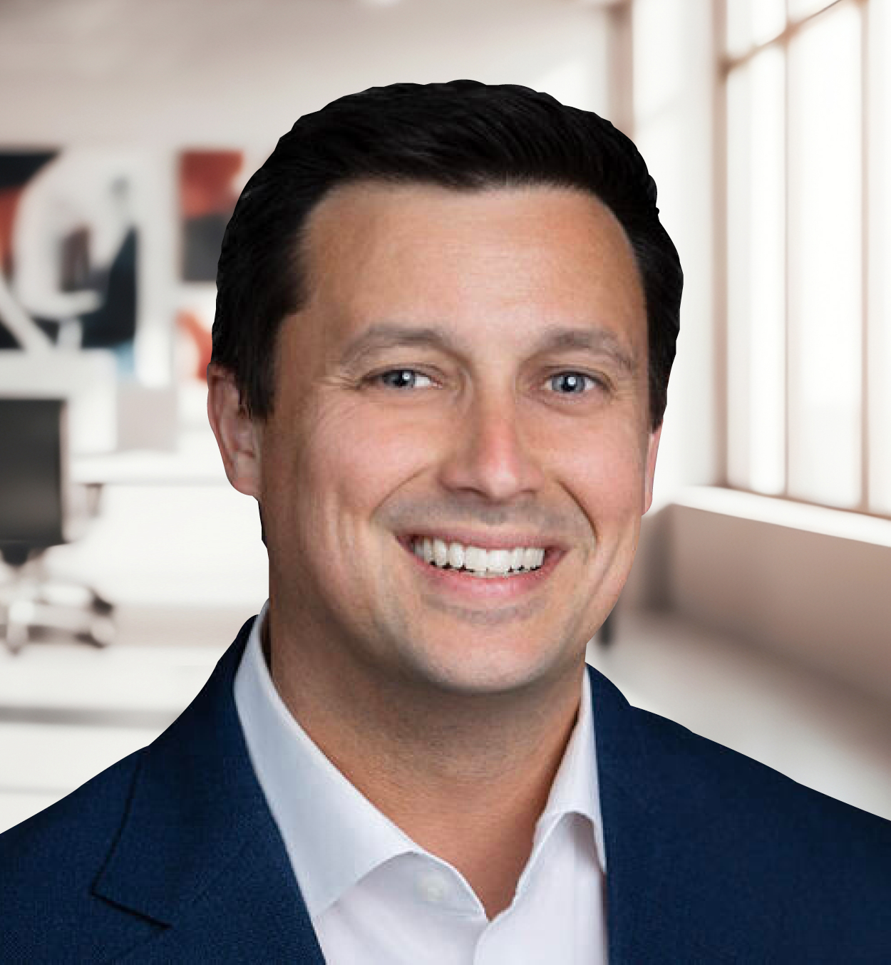 A man with dark hair, blue eyes, wearing a navy suit and white shirt, smiling in an office setting with large windows and office furniture in the background.