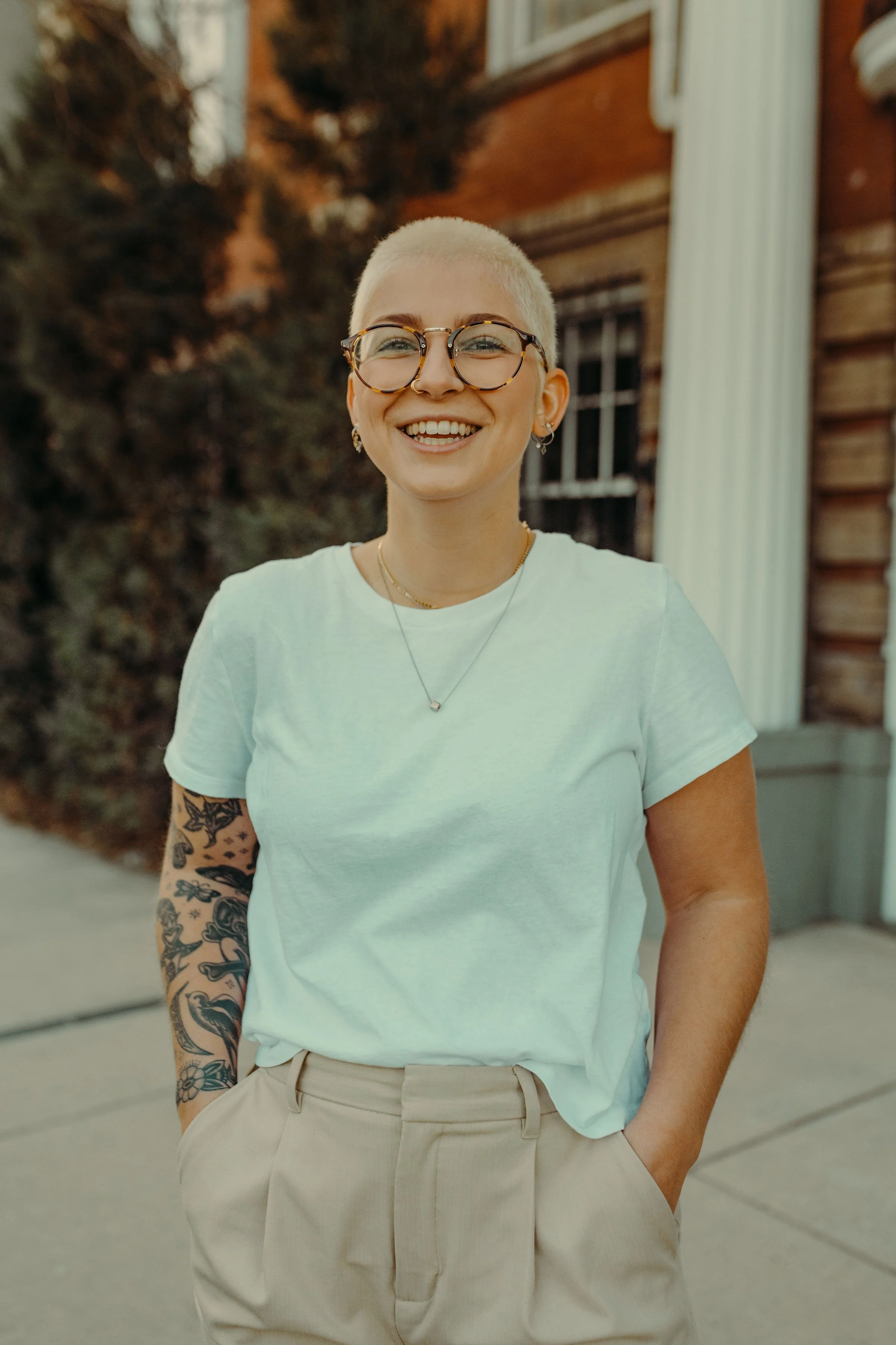 A woman with a platinum blonde buzz cut hairstyle, wearing round glasses, a white t-shirt, beige trousers, and jewelry, smiling outdoors with a blurred background of trees and a brick building.