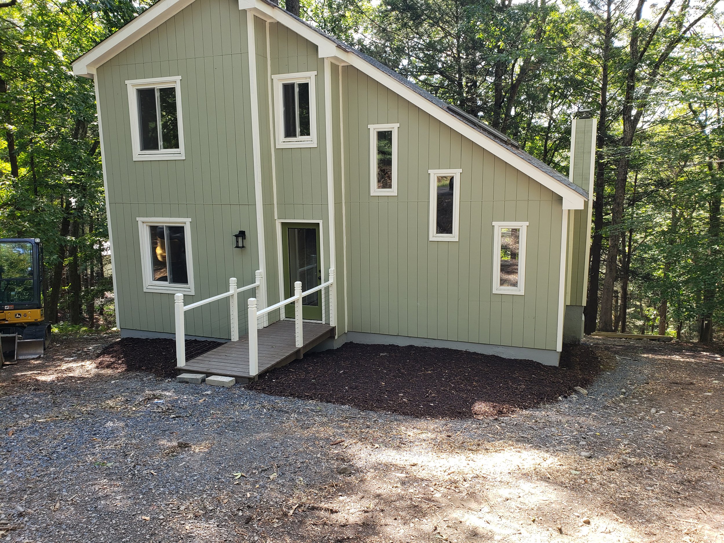 A modern two-story house with light green siding, white trim, and multiple narrow windows situated in a wooded area. The house has a small wooden ramp leading to the front door.
