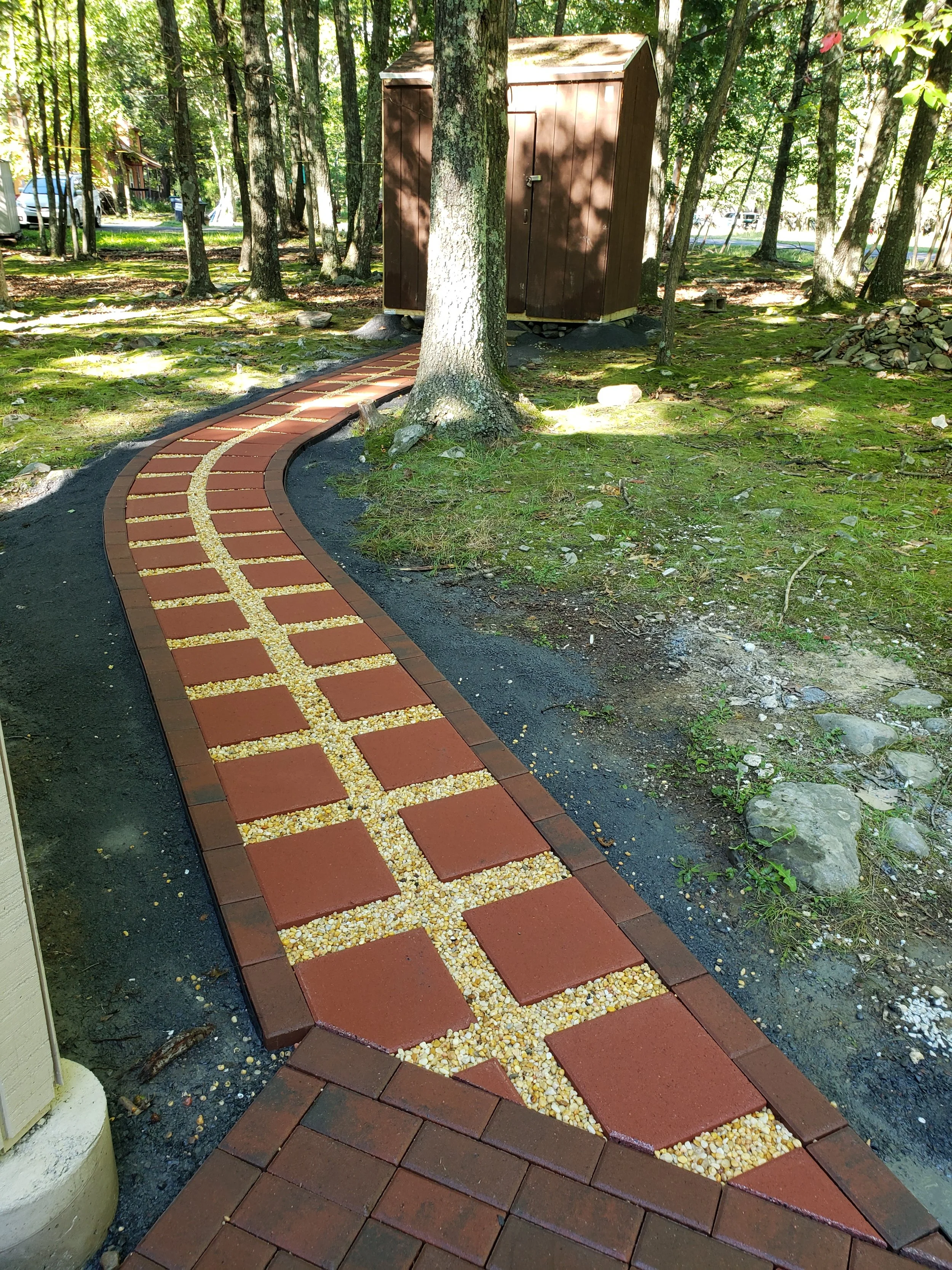 Colorful brick pathway winding through a forested area with trees and moss-covered ground, leading to a small brown shed.