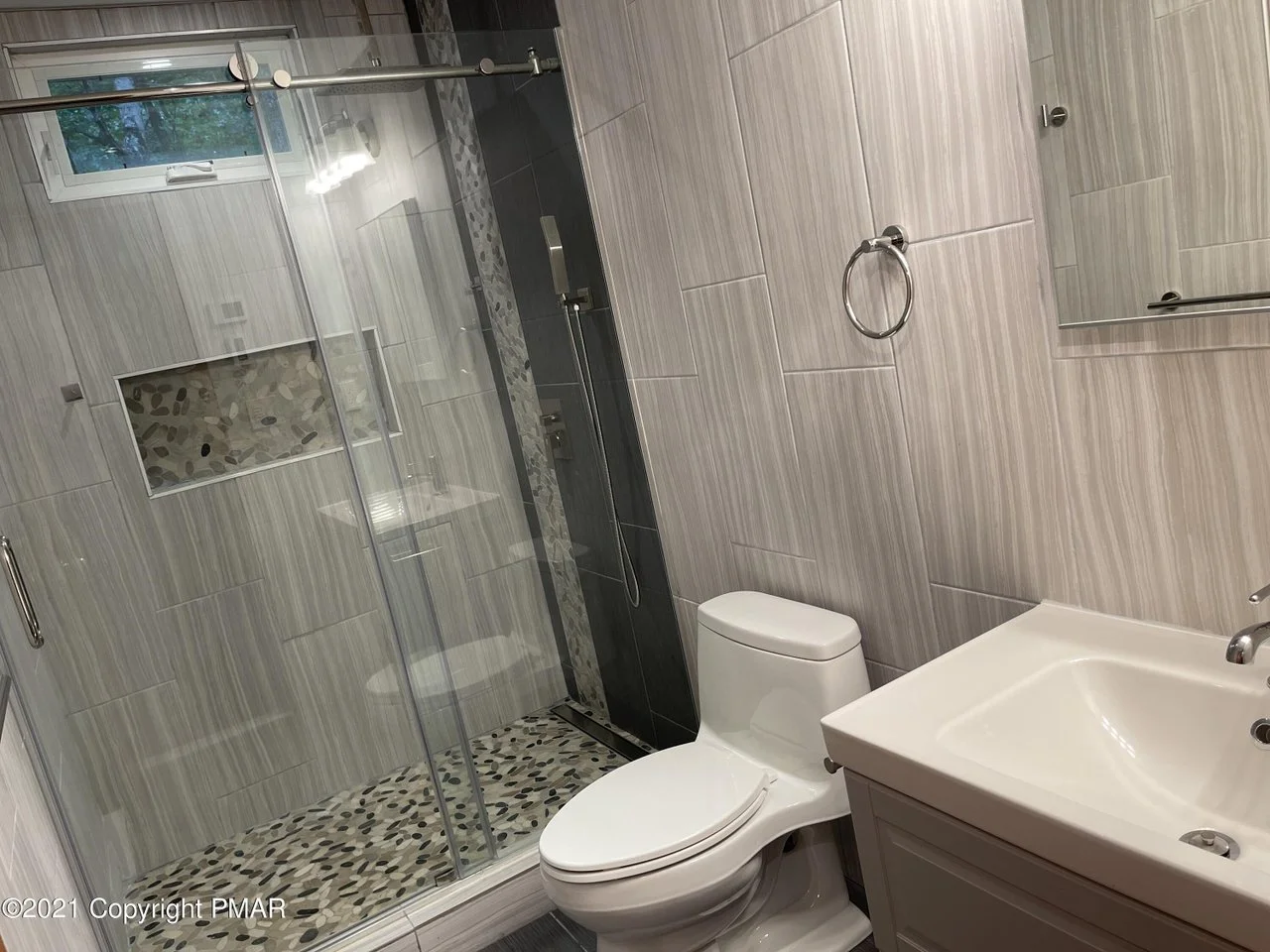 Modern bathroom with a glass shower enclosure, pebble tile floor in the shower, beige wall tiles, toilet, sink, and mirror.