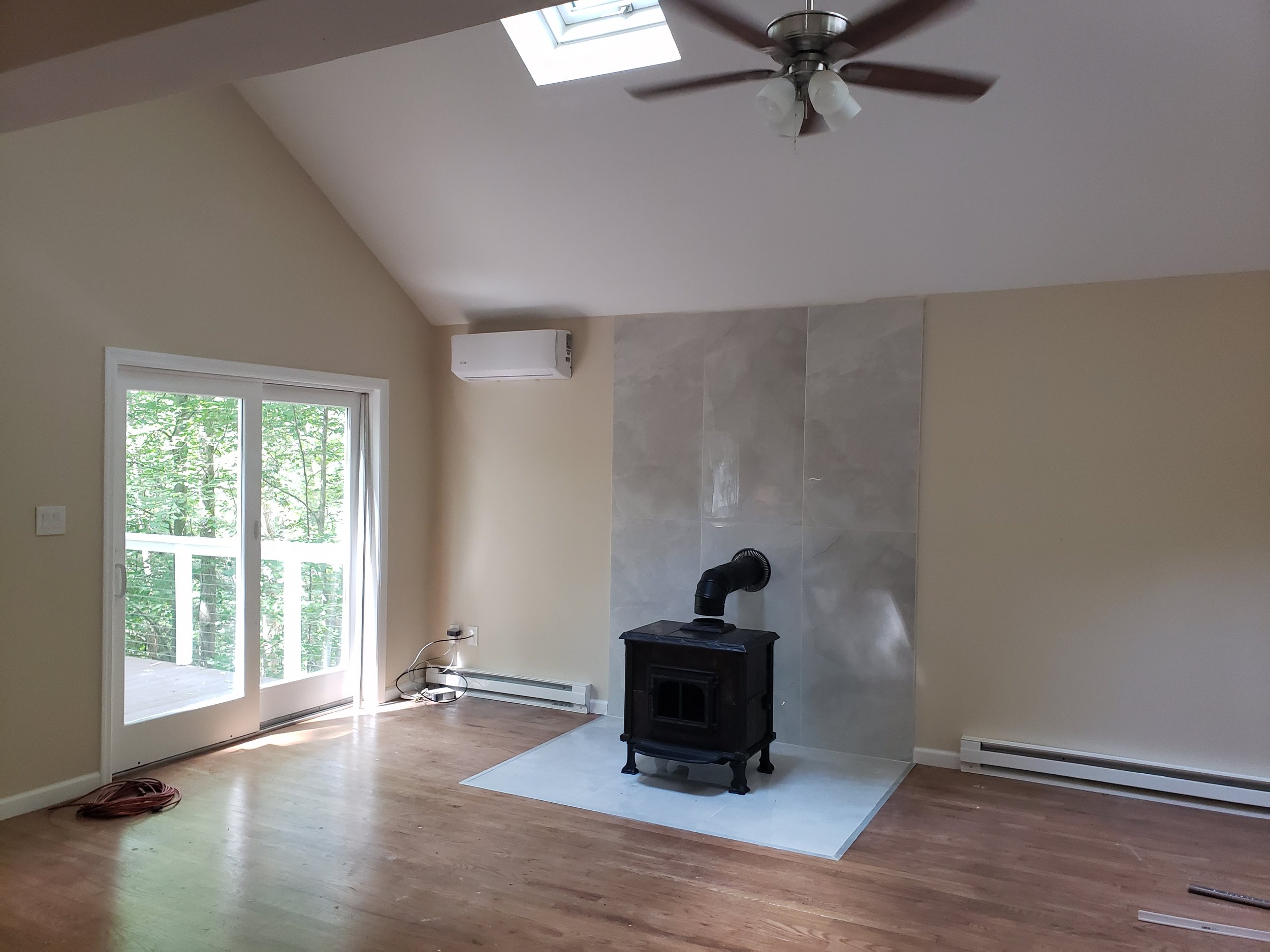 Unfurnished living room with a wood stove on a tiled hearth, a ceiling fan, and a sliding glass door leading to a balcony showing trees outside. There is an air conditioning unit on the wall and some electrical cords on the floor.