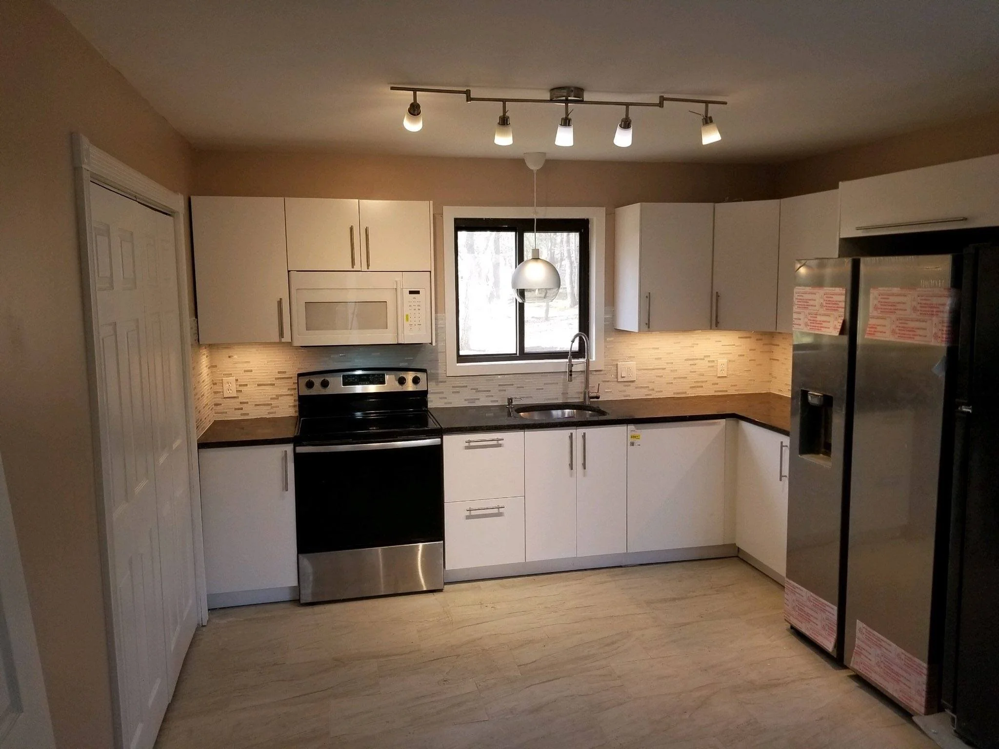 Modern kitchen with white cabinets, black countertops, stainless steel appliances including a refrigerator and oven, a microwave, backsplash with beige tiles, a window above the sink, and track lighting on the ceiling.