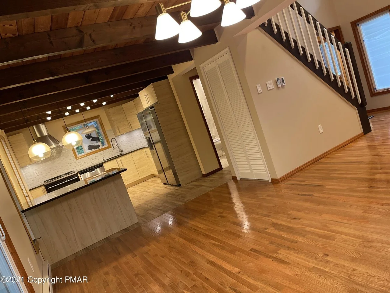 Modern kitchen with wooden cabinets, a kitchen island, three pendant lights, and stainless steel appliances, including a refrigerator, set against a white textured backsplash and a window.