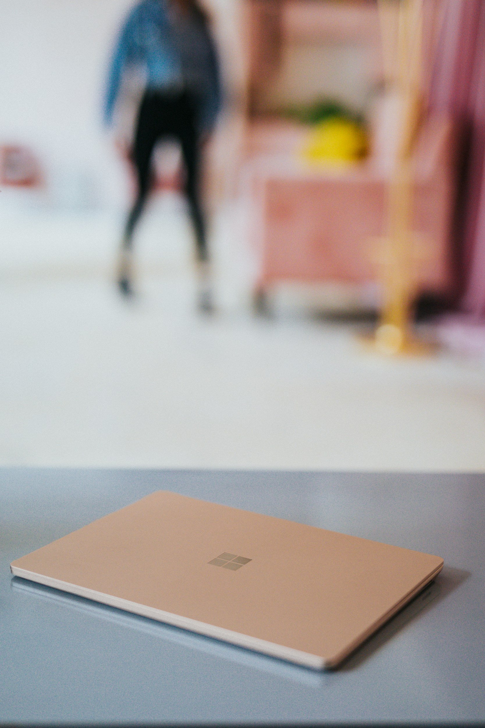 A closed peach-colored Microsoft Surface laptop resting on a white desk in the foreground, with a blurred background featuring a person standing in a room with pink and yellow furniture.