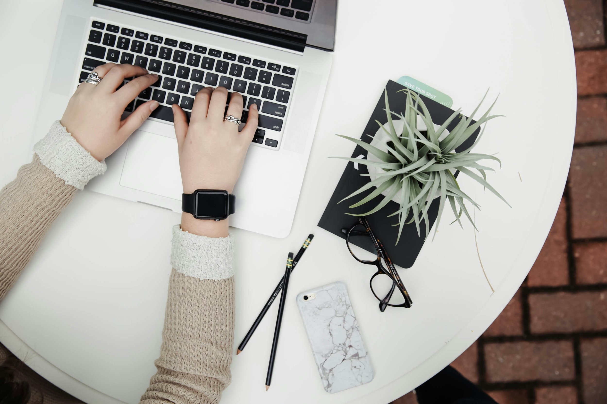 Person typing on a laptop at a white desk, with a notebook, plant, glasses, pencils, and a marble phone case nearby.