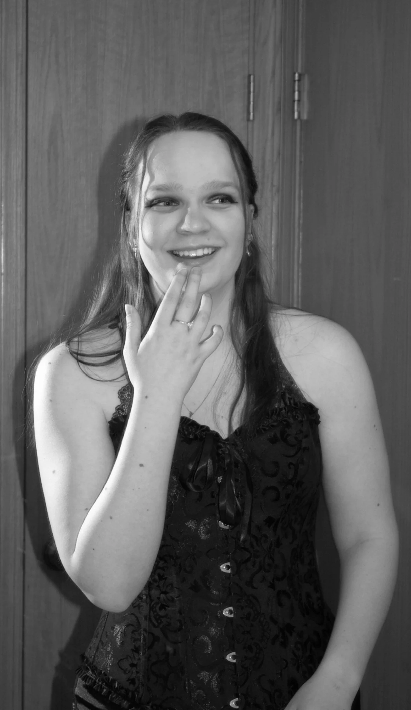 A smiling young woman in a black, sleeveless, patterned dress with a lace trim, touching her face with her left hand, standing in front of a wooden door.