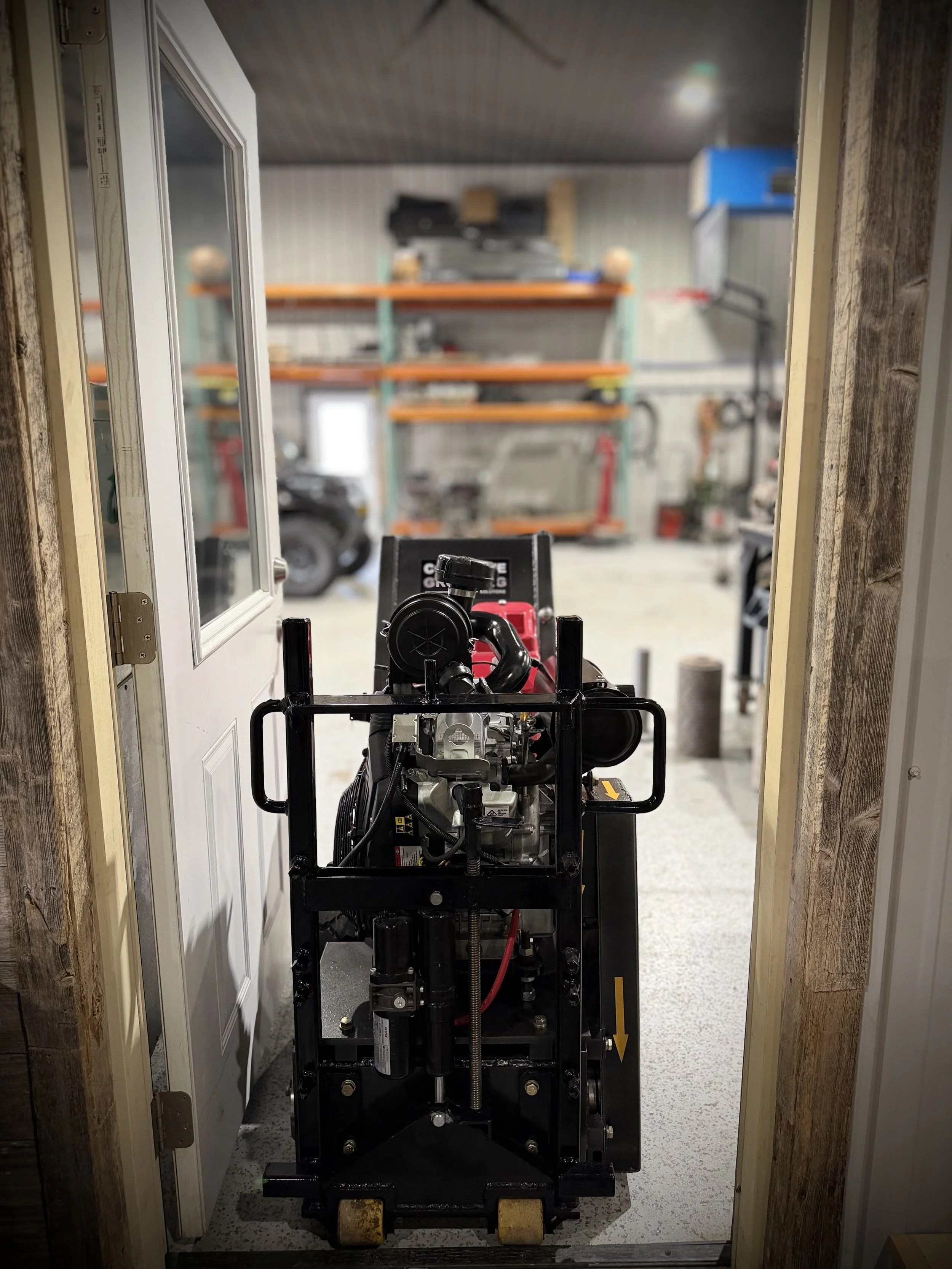A mechanical device or piece of equipment is positioned in a doorway, with a garage or workshop area in the background. The device appears to be a machine or engine with various components and parts.