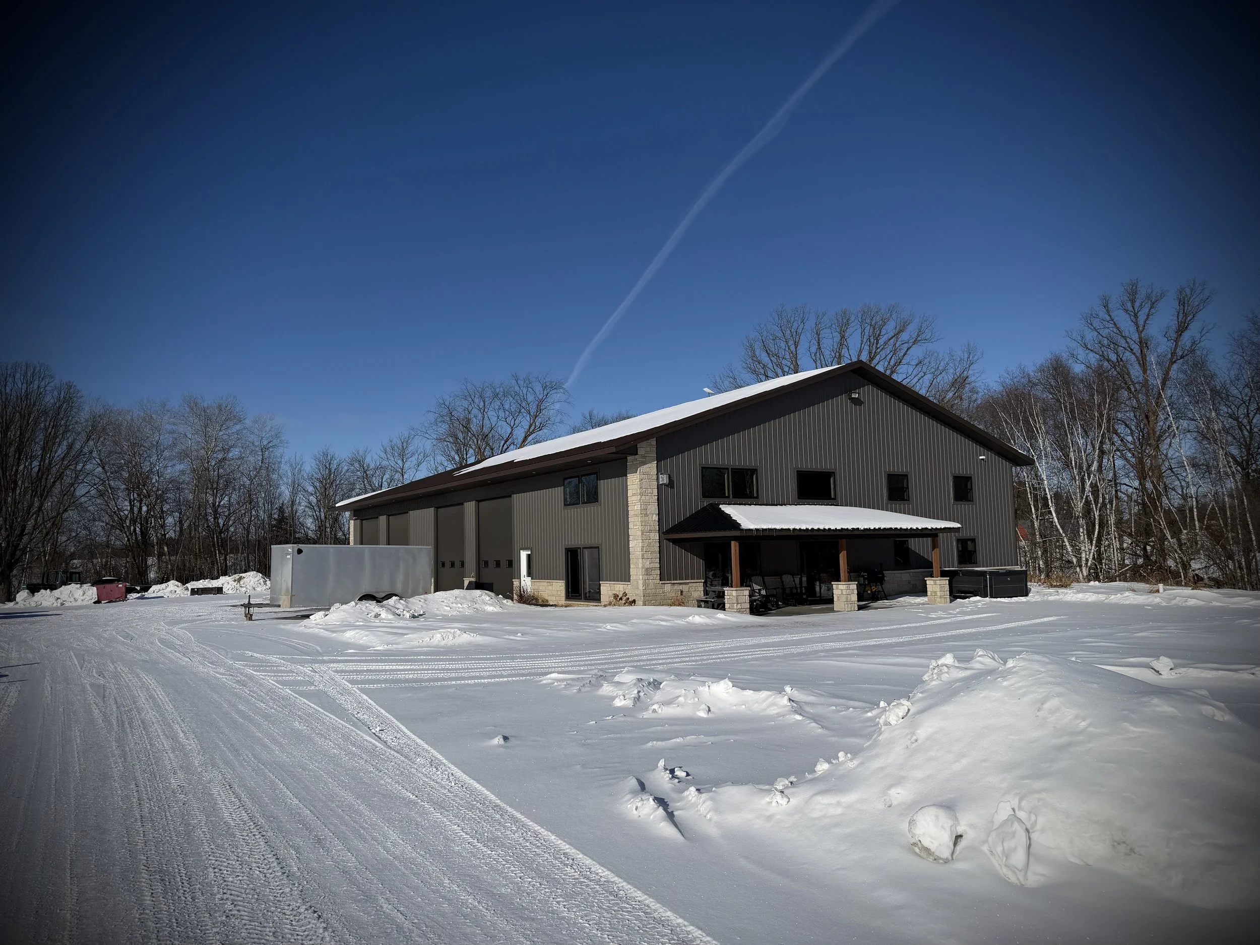 A large modern building with dark siding and a sloped roof, situated in a snowy landscape with tire tracks, leafless trees in the background, and a clear blue sky with a contrail.