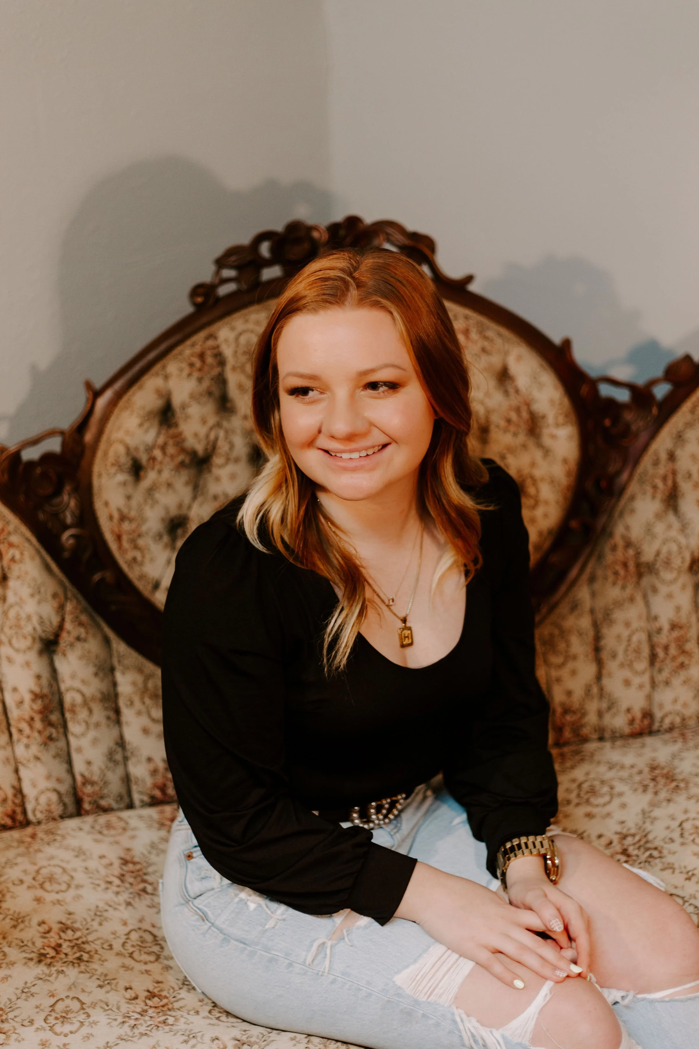 A young woman with red hair sitting on a vintage floral sofa, smiling and looking to her right, wearing a black top, ripped jeans, a gold watch, and necklaces.