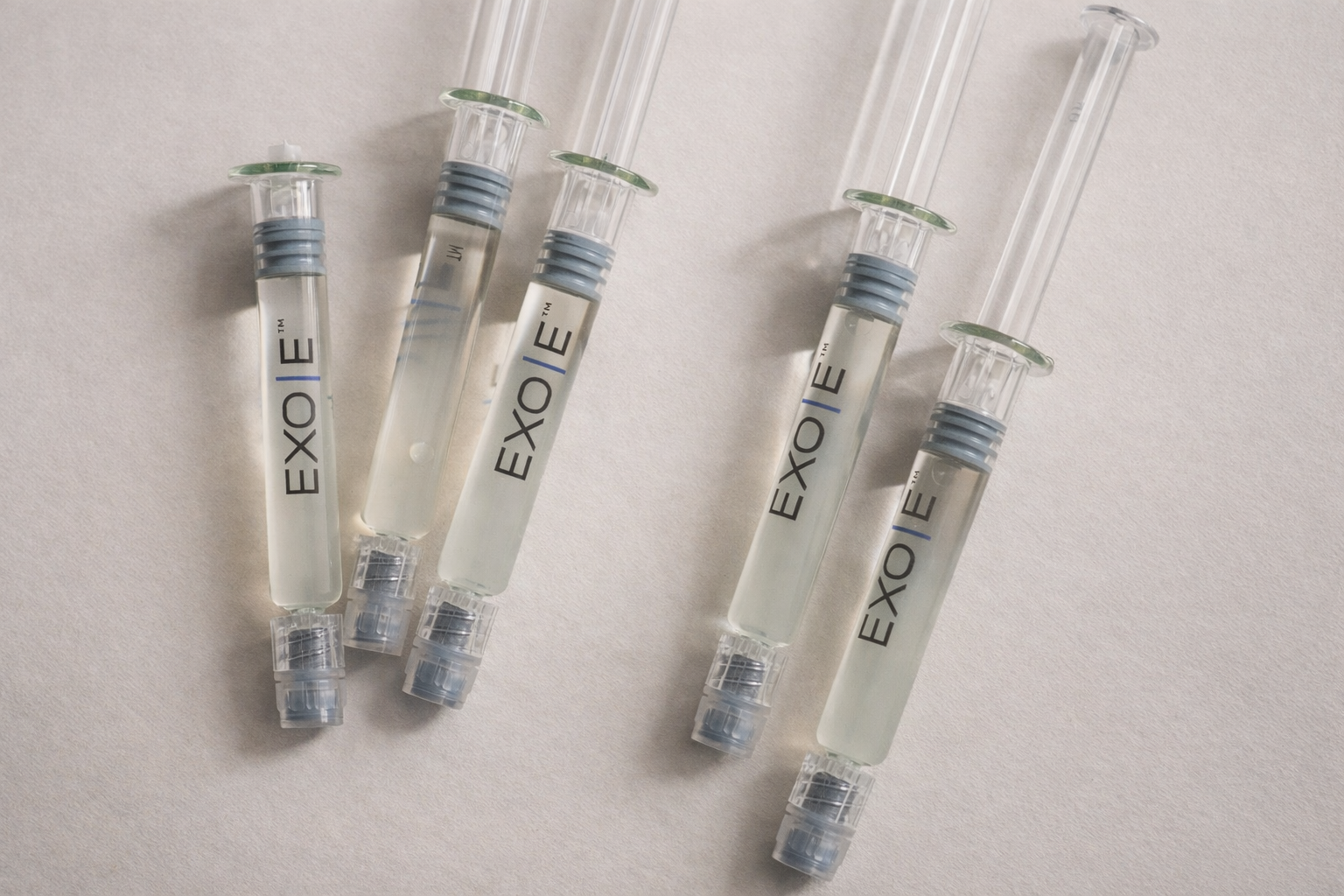 Six syringes with clear liquid and clear plastic needles on a light surface.