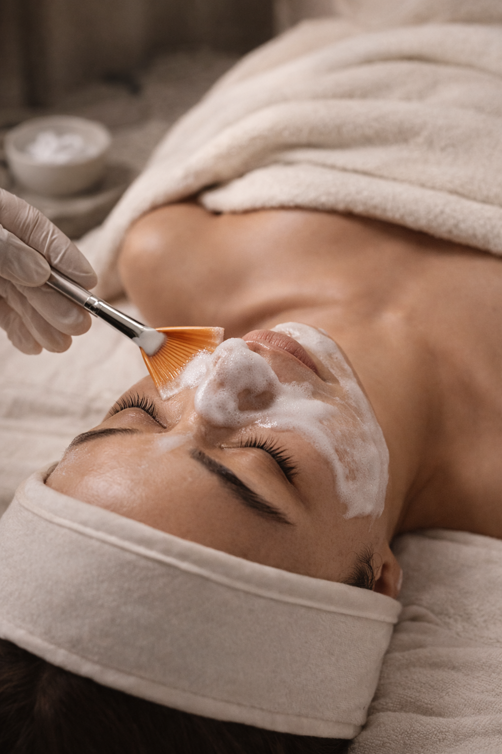 A woman receiving a facial treatment with foam, while a esthetician applies a facial mask using a brush.