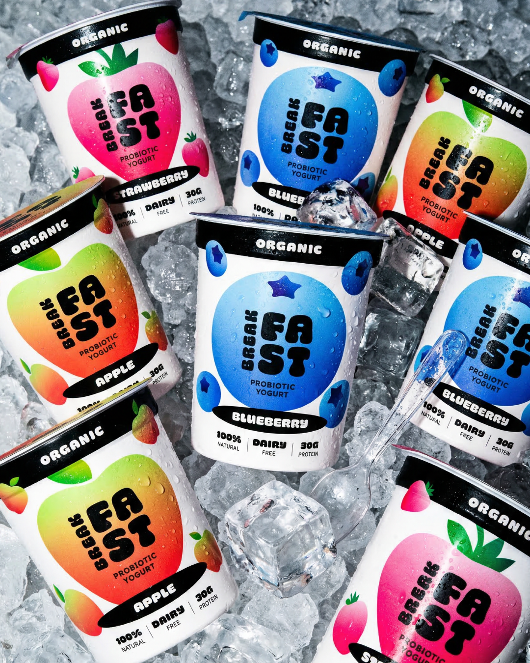 Multiple cans of Breakfast Fast probiotic yogurt in apple, blueberry, strawberry, and peach flavors, placed on ice with ice cubes and a plastic spoon.