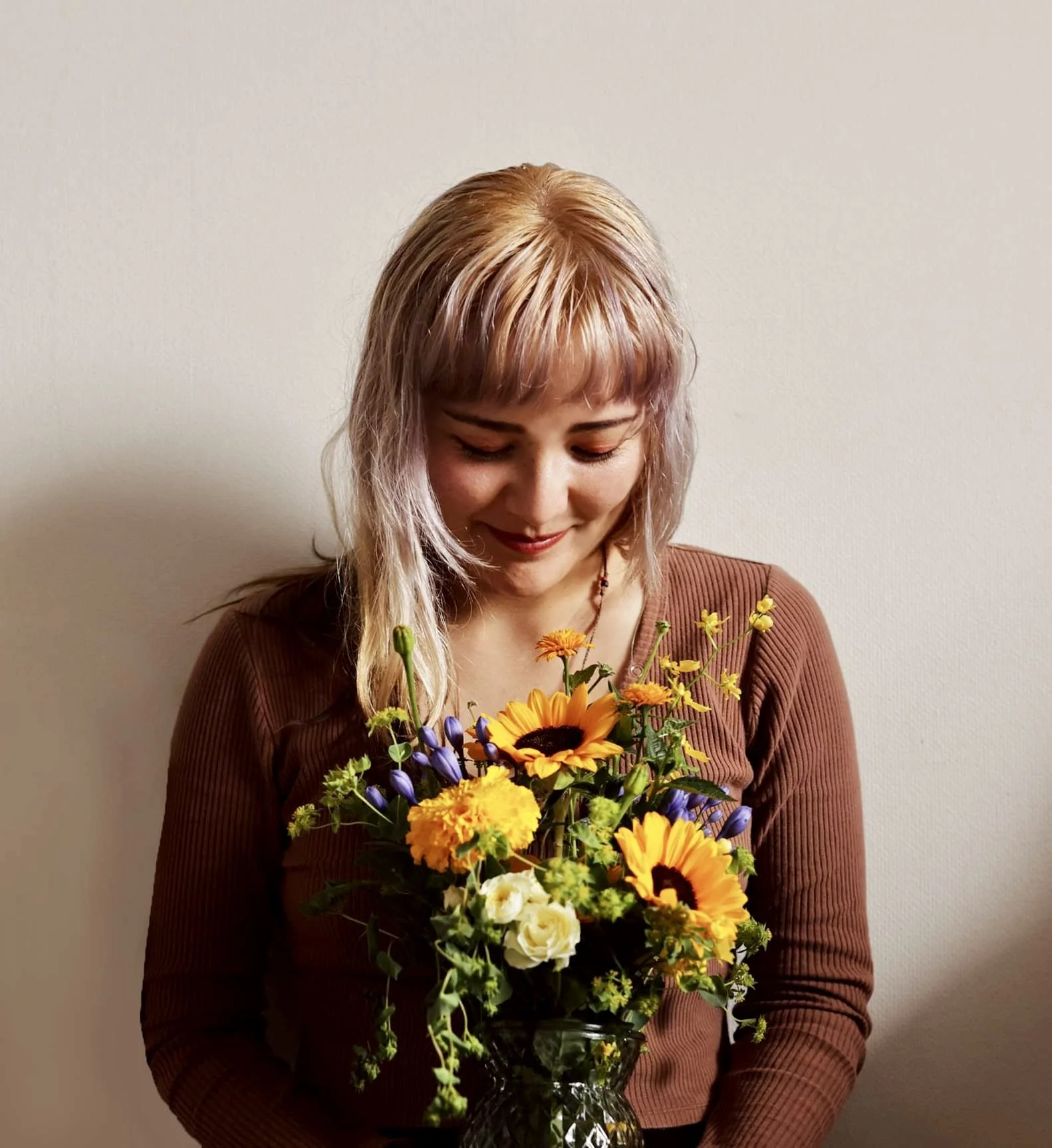 A woman with light hair and a warm smile is holding a bouquet of colorful flowers, including sunflowers, yellow, white, and purple blooms, in a dark vase against a plain light-colored wall.