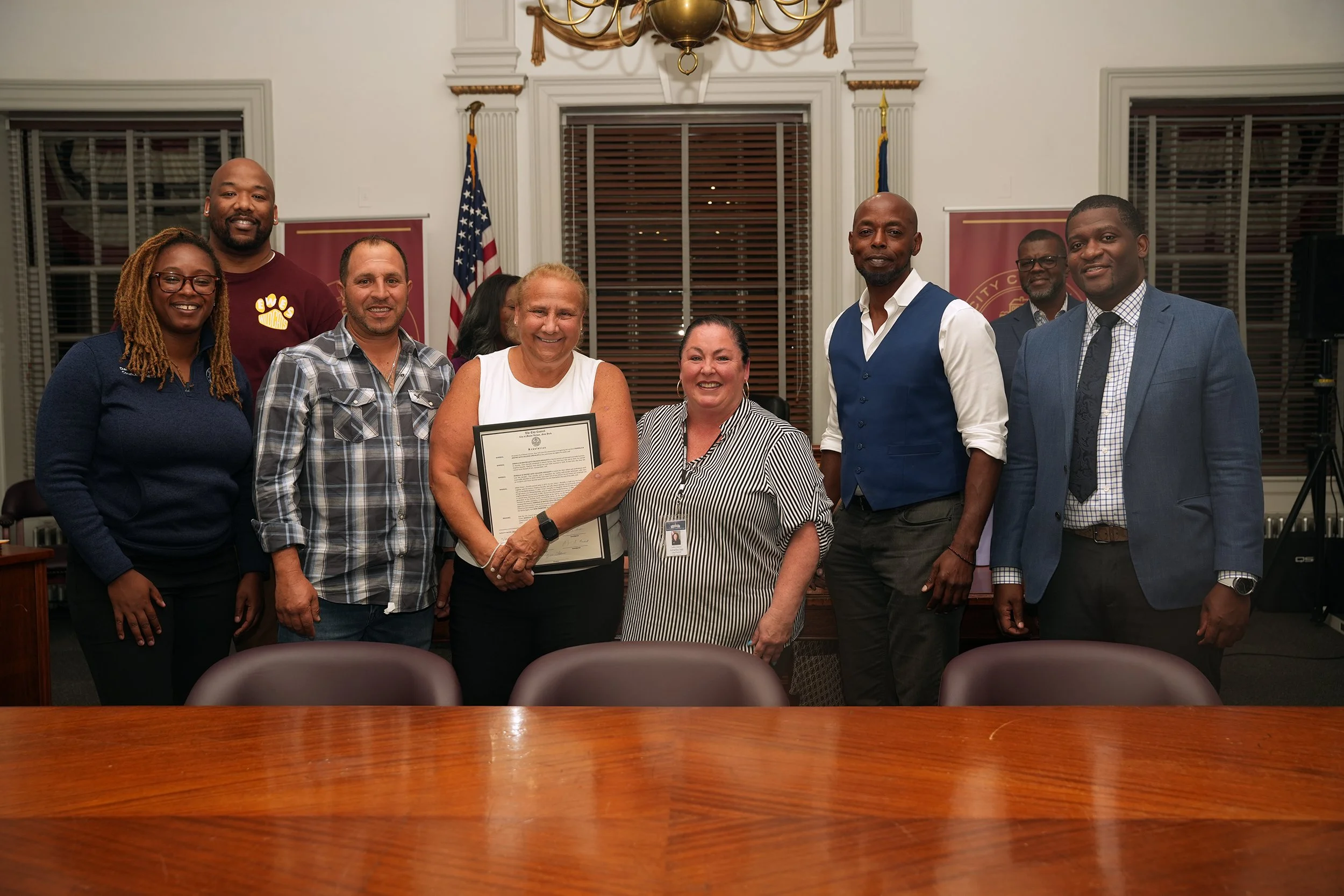A group of nine diverse adults standing in a conference room, posing for a photo. The woman in the center is holding a framed certificate, with American flags and wooden blinds in the background.