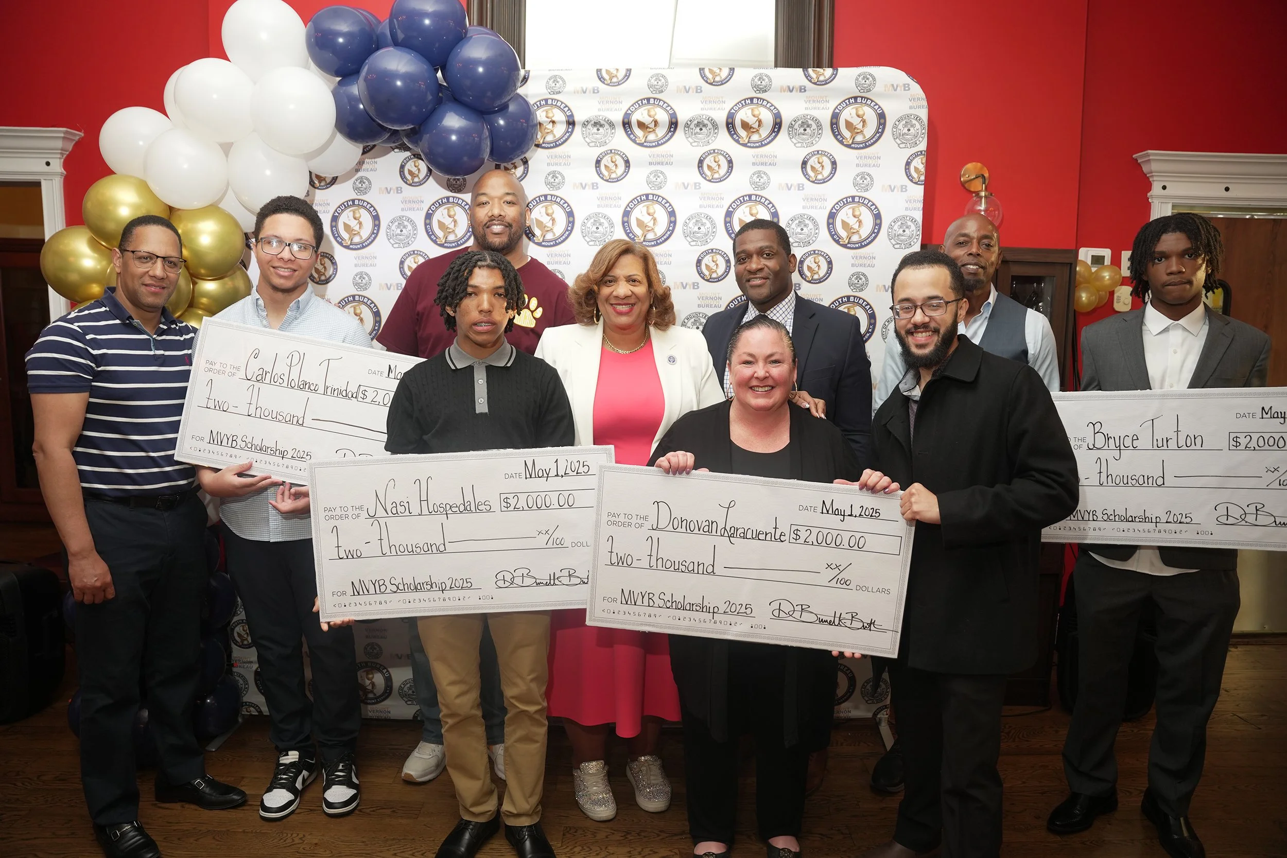 Group of people holding oversized checks at a scholarship event, with balloons and a backdrop displaying a logo in the background.