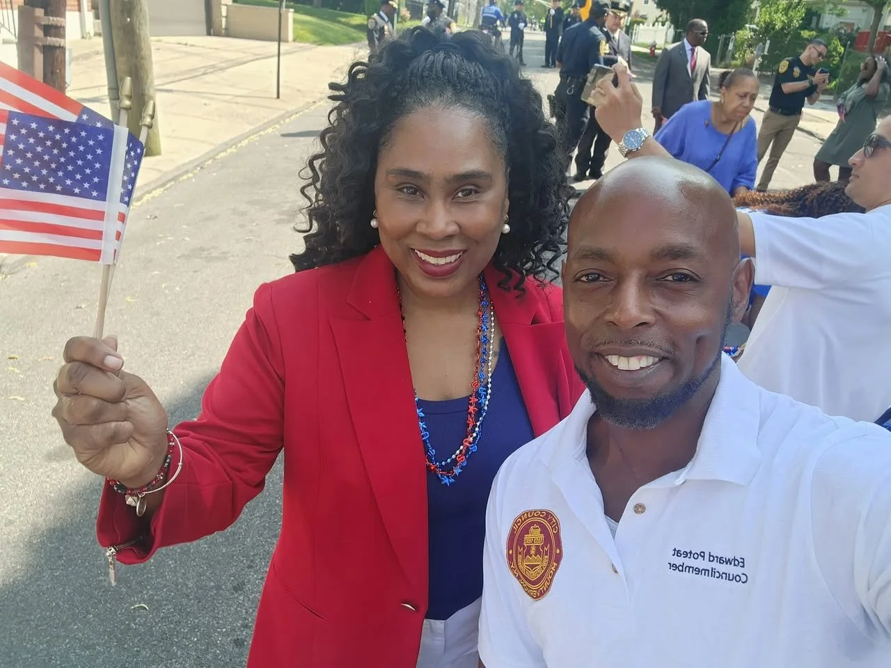 A woman in a red blazer holding an American flag taking a selfie with a man in a white polo shirt at a public event, with other people in the background.