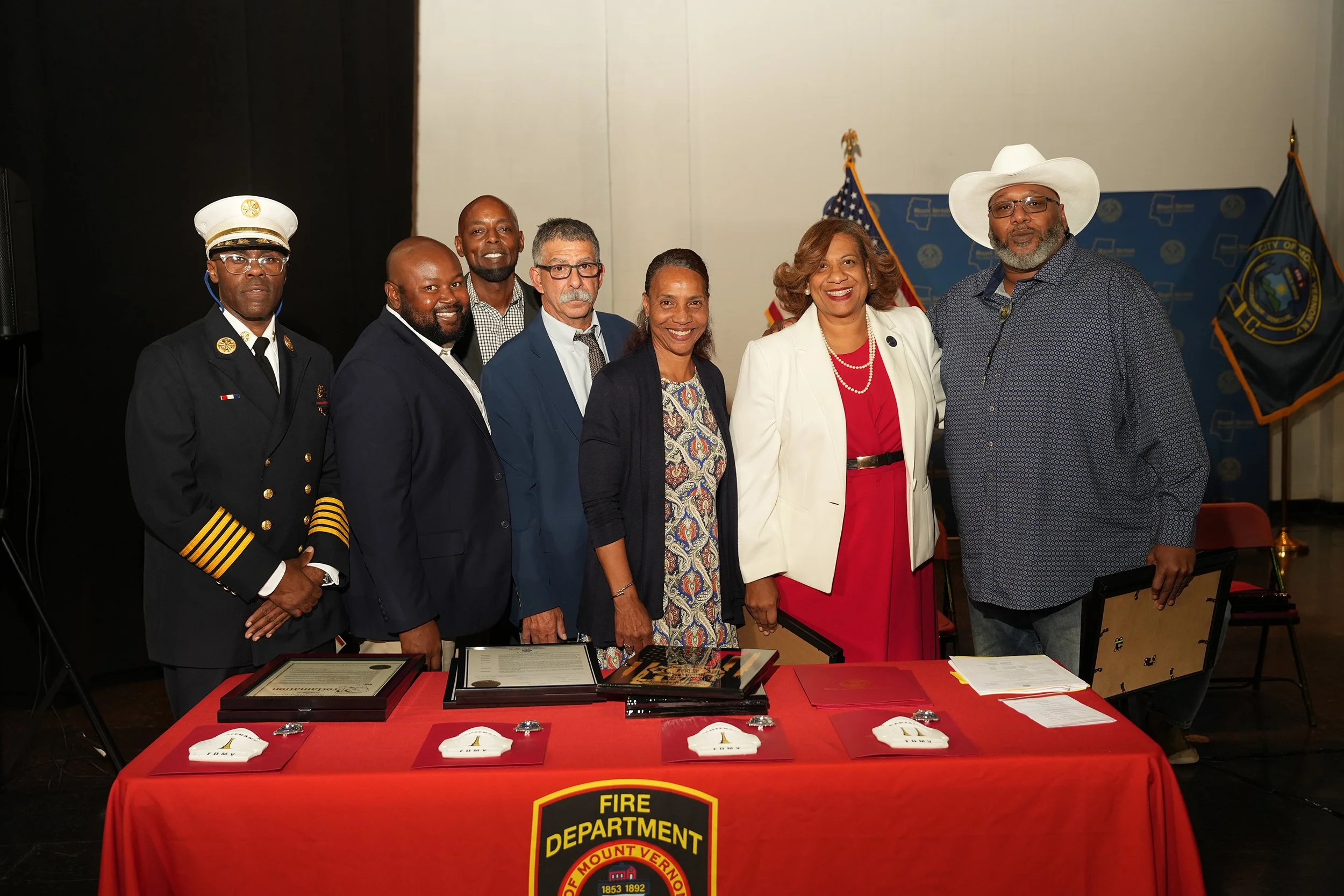 Group of eight diverse people standing behind a table with documents and awards, in a room with a fire department flag and American flag in the background.