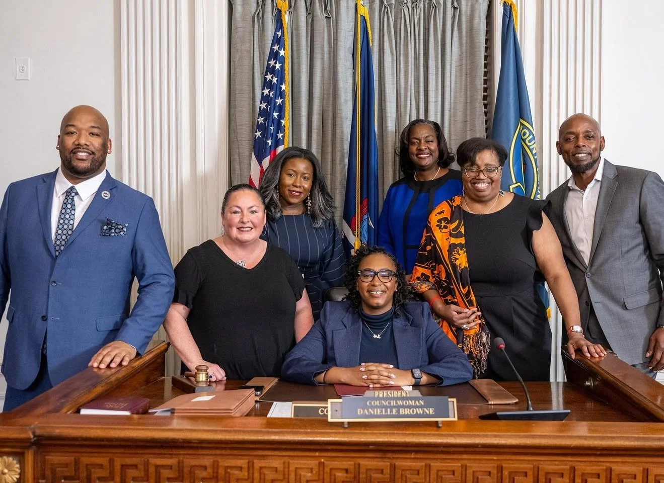 Group of eight diverse people, six women and two men, posing in a formal setting with flags behind them. One woman seated at a desk in front holds a nameplate reading "Councilwoman Danielle Browne."