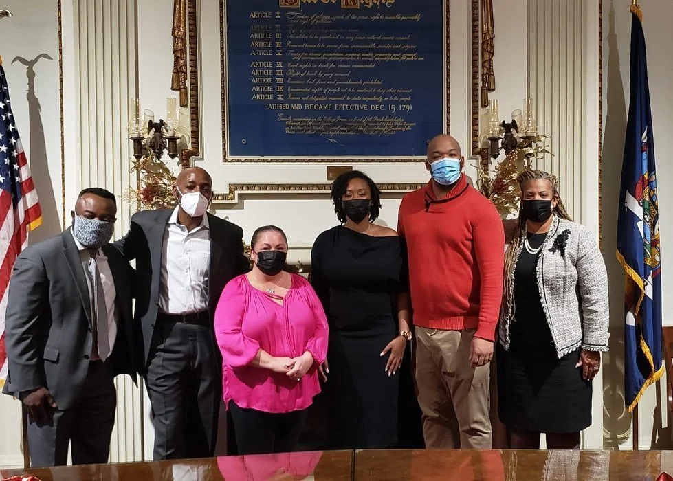 Six people wearing masks standing in a room with flags and a framed document or artwork behind them.