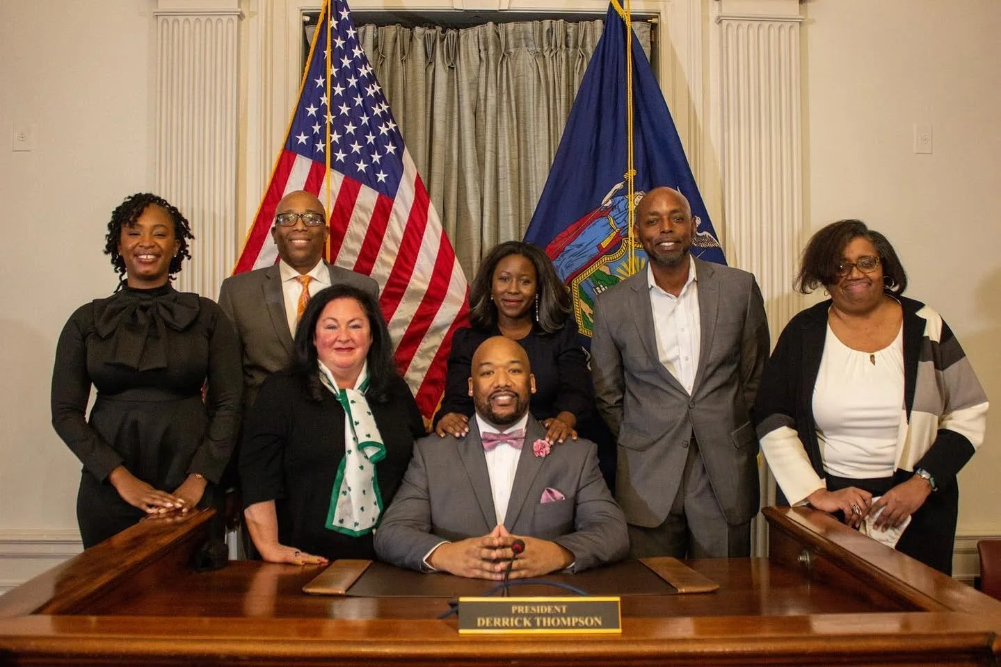 Group photo of eight people, including President Derrick Thompson seated at a wooden desk with a nameplate, in a formal setting with American and state flags in the background.
