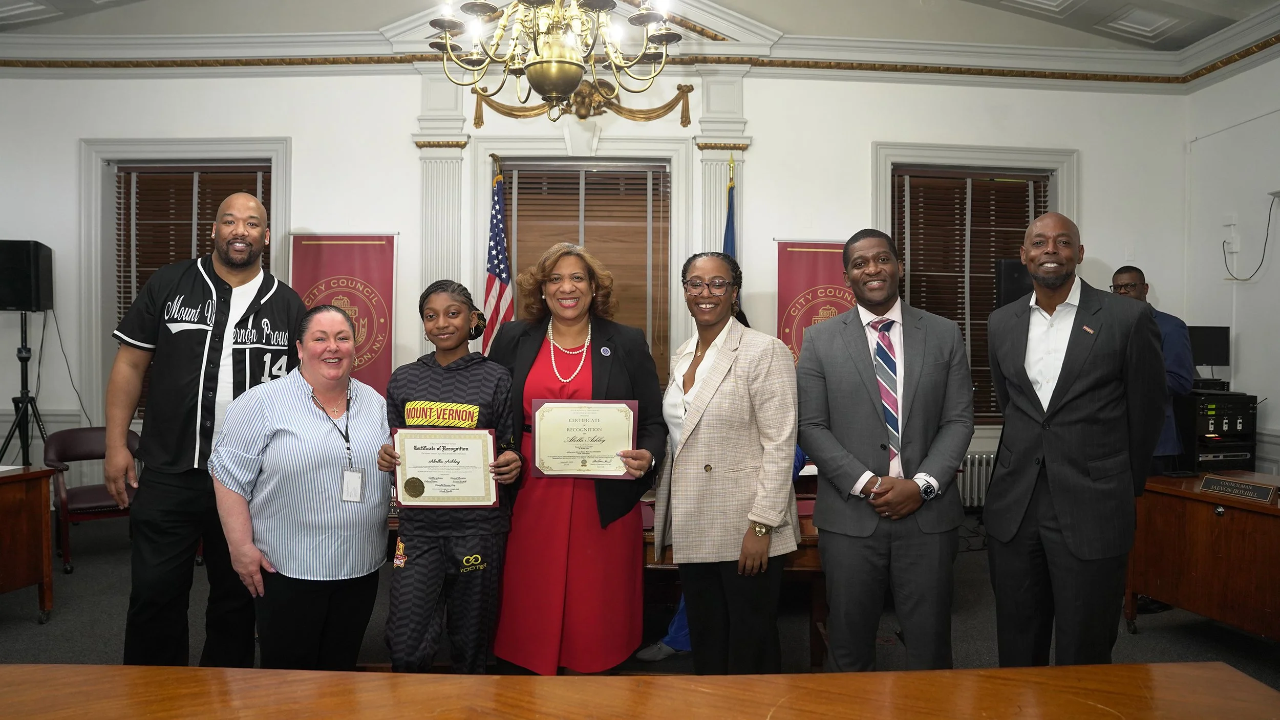 A group of seven people standing in a room with wood-paneled windows and flags, some holding certificates, celebrating an award ceremony.