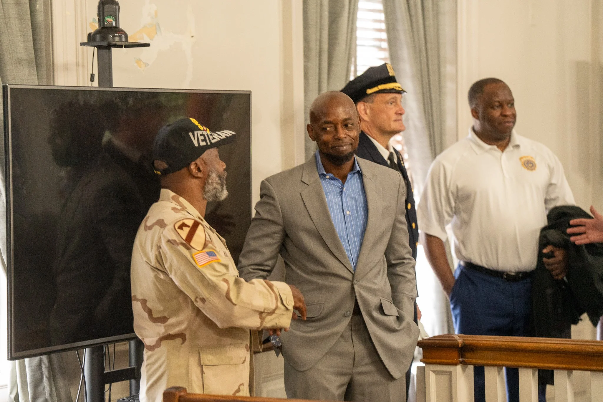 Group of men, including a man in military uniform and a man in a gray suit, standing in a room with a television and curtains.