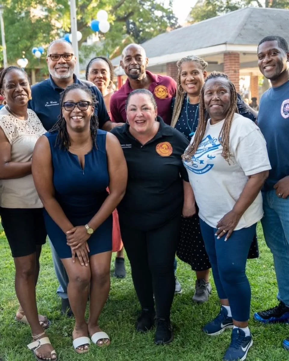 A group of ten diverse adults smiling together outdoors at a park or outdoor event, with a pavilion and trees in the background.