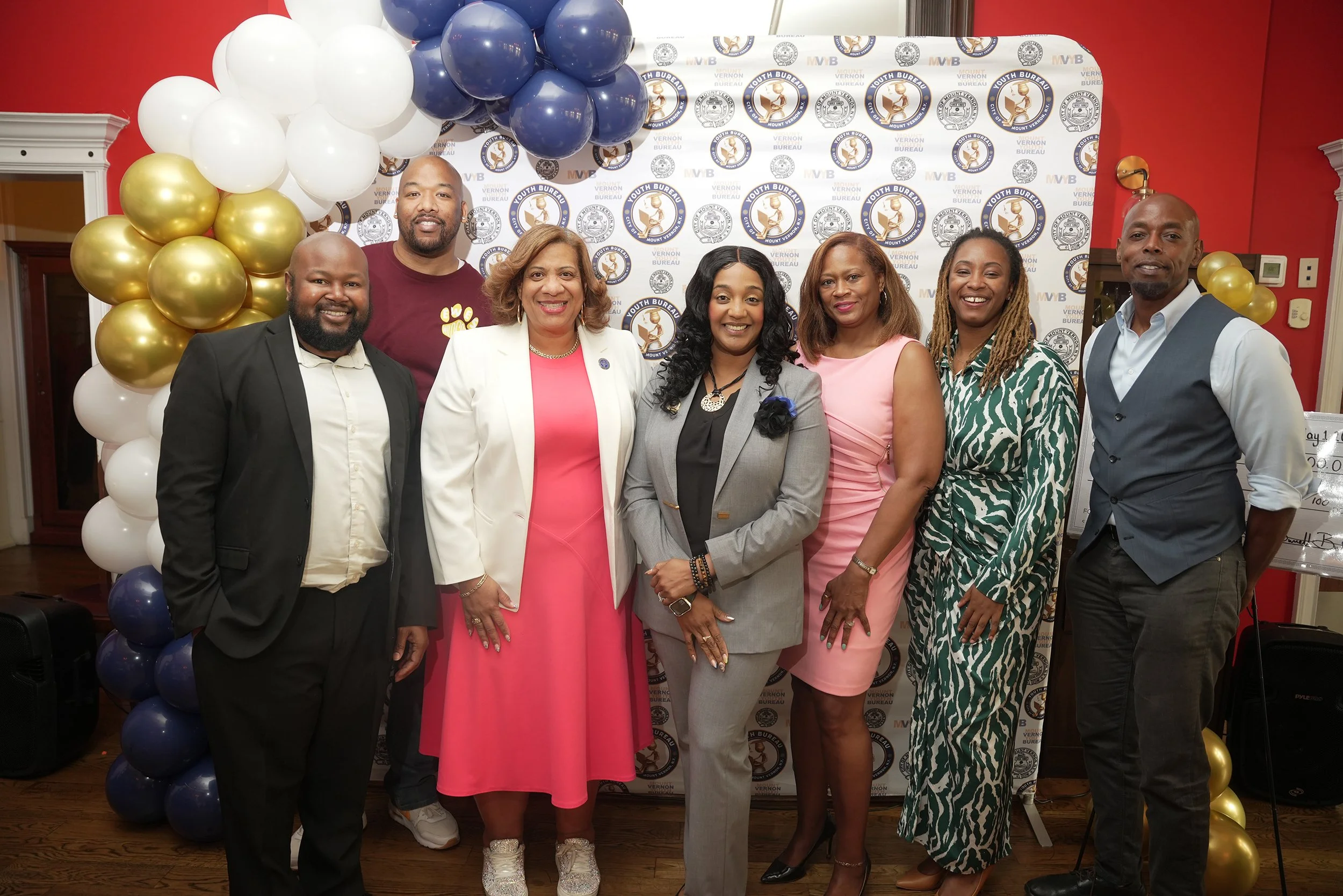 Group of eight diverse adults in formal attire standing together in front of a backdrop with logos, surrounded by balloon decorations, at a formal event.