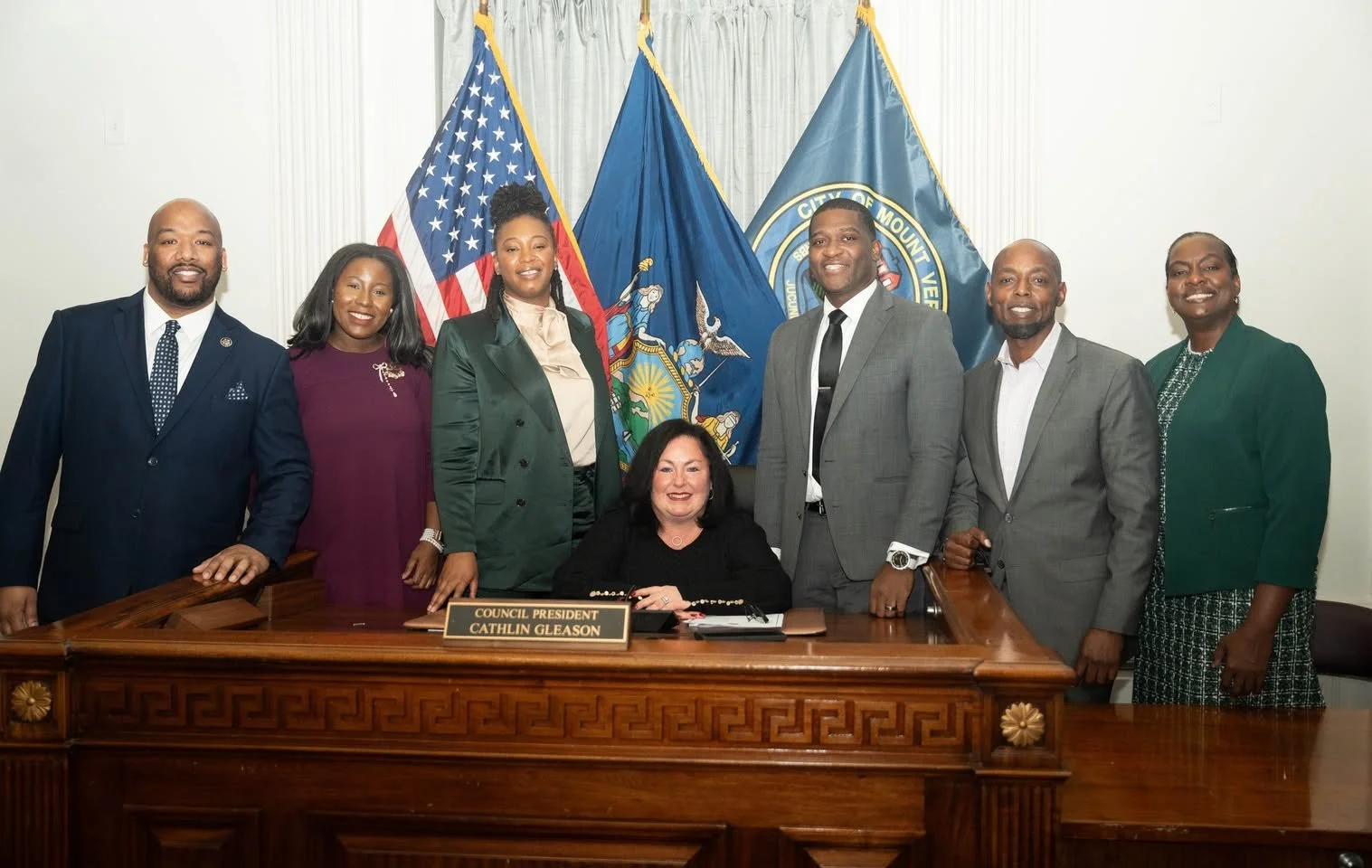 A group of eight diverse people, six men and two women, standing and sitting behind a wooden desk with a nameplate that reads 'Council President Cathlin Gleason', in front of flags including the American flag, and city flags, in a government setting.
