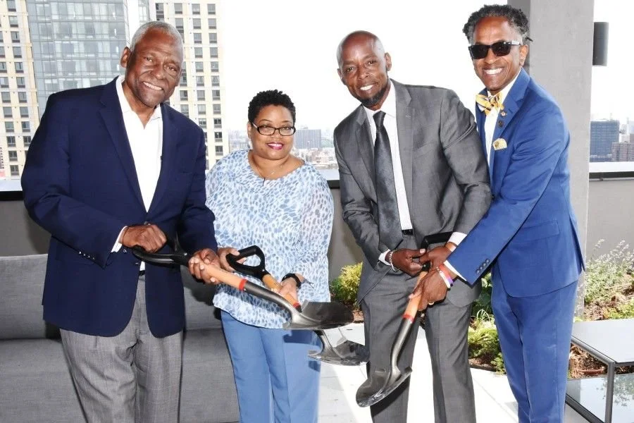 Four professionally dressed people, including three men and one woman, holding shovels in a city rooftop setting during the daytime, smiling at the camera.