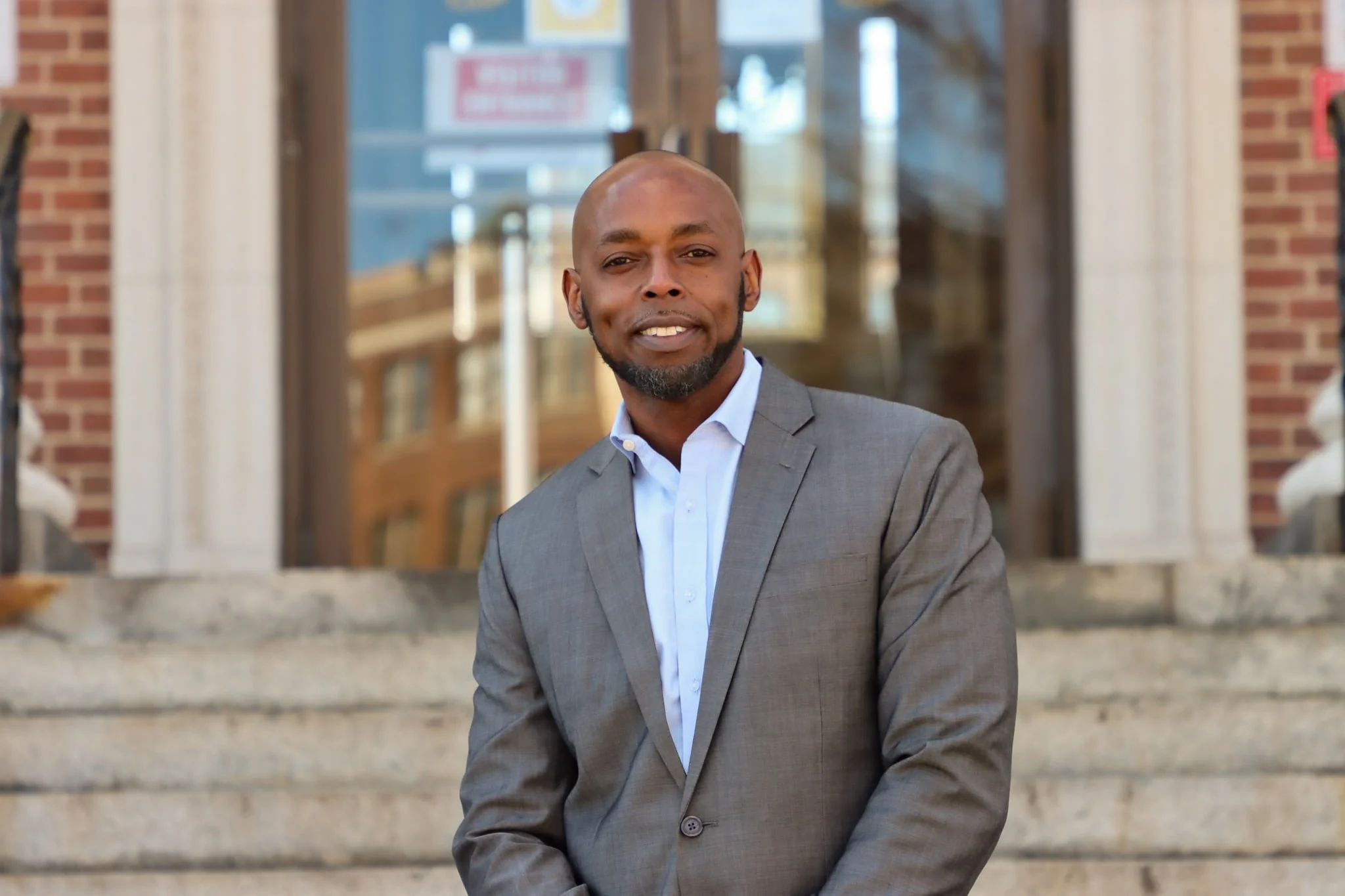 A man in a gray suit and white shirt standing outside in front of a brick building with a window.