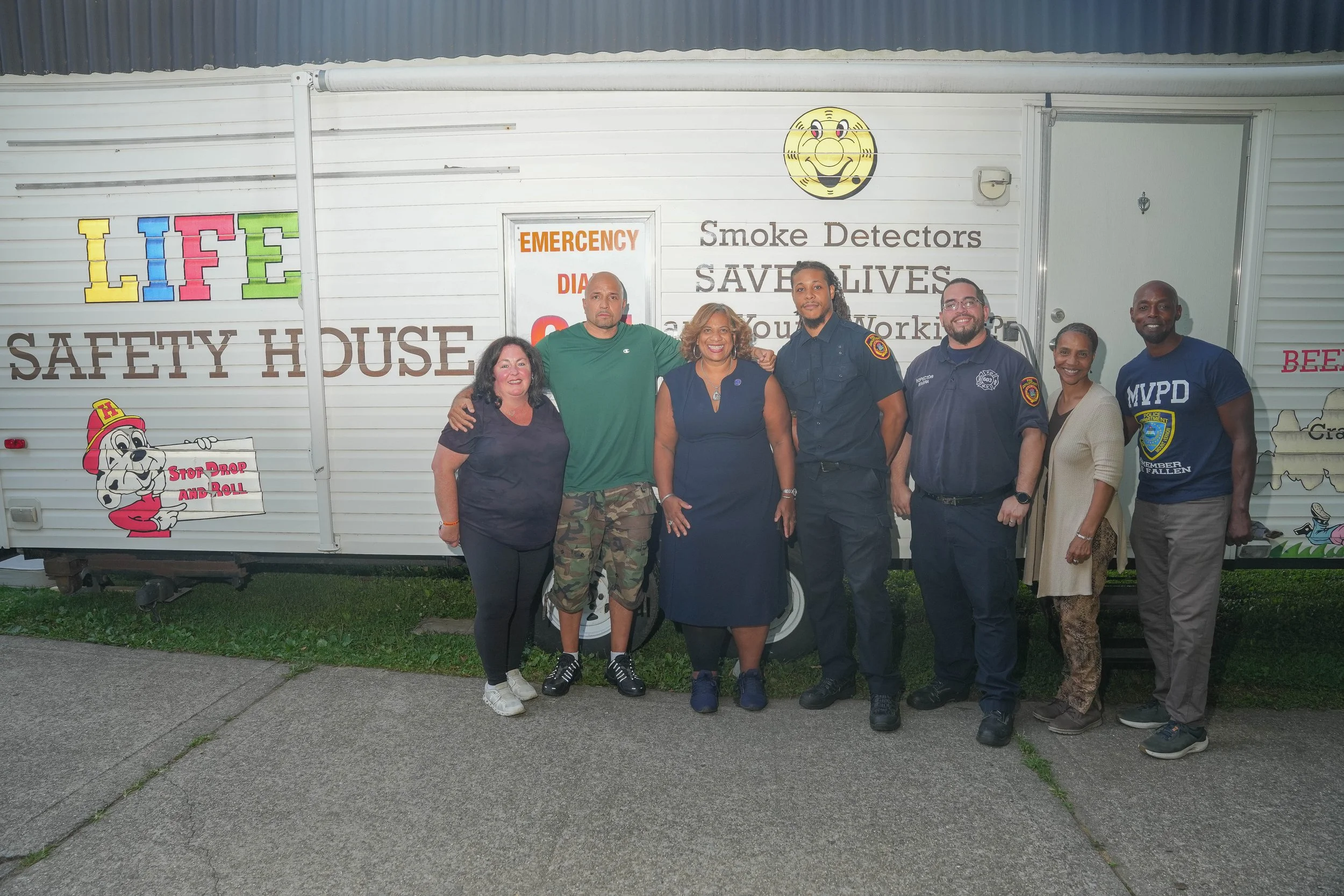 Group of seven people standing in front of a safety house with various signs and slogans related to fire safety and community services, including a cartoon dog holding a sign that says 'Stop Drop and Roll'.