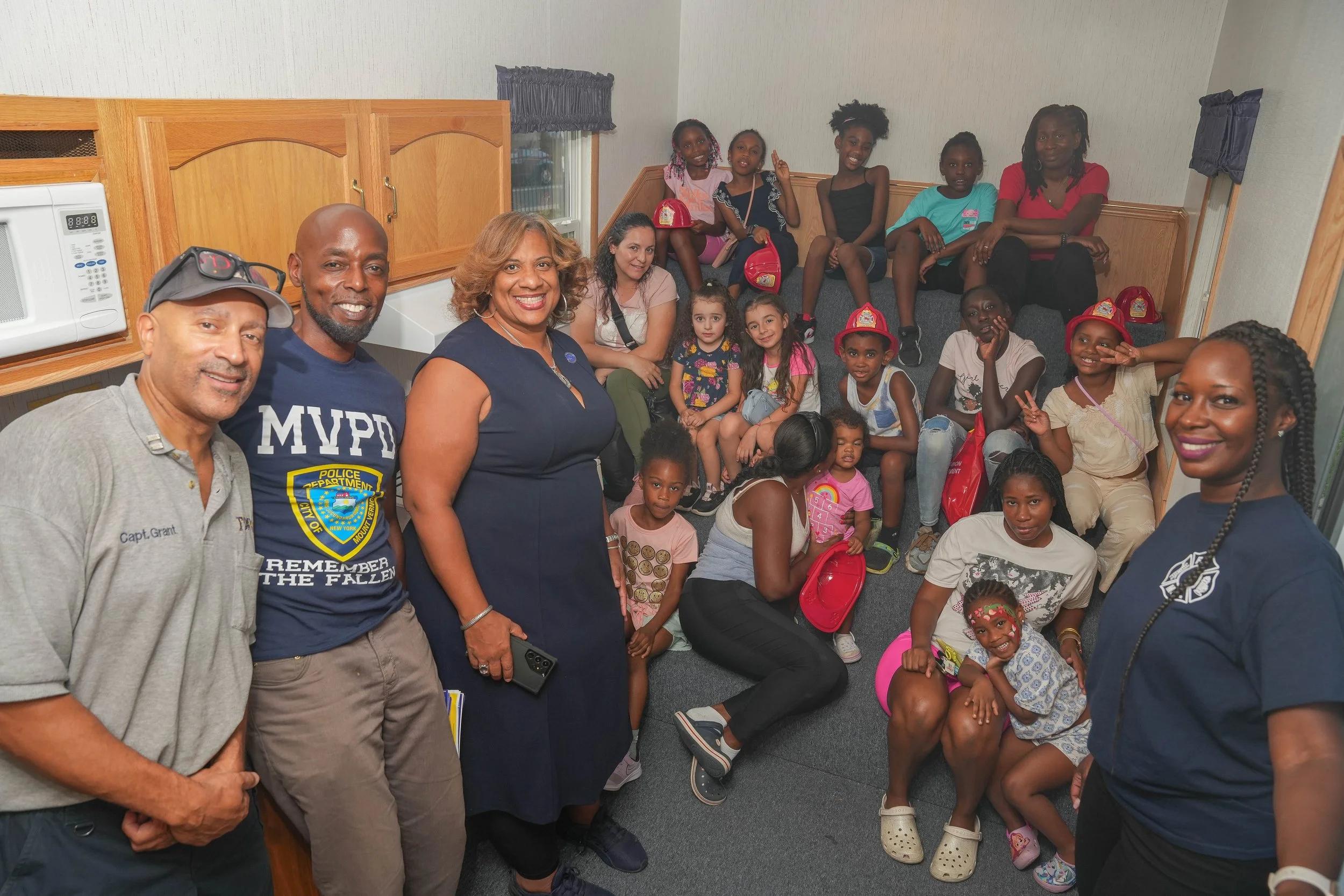 Group of children and adults gathered in a small room, smiling for a photo. Some children are wearing red firefighter helmets, and the group appears to be participating in a community event or visit.