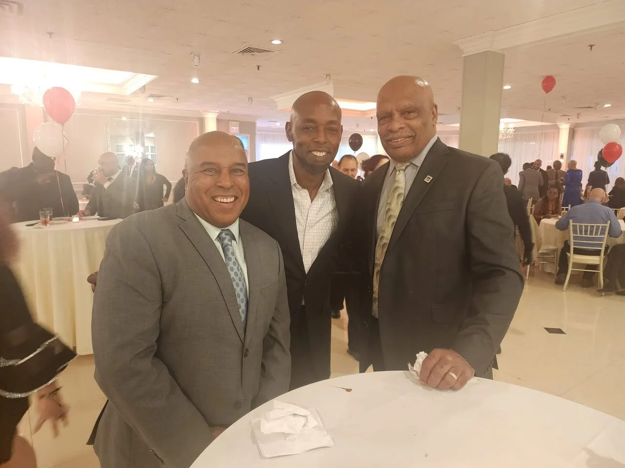 Three men in suits standing together at an indoor event, smiling at the camera, with balloons and other people in the background.