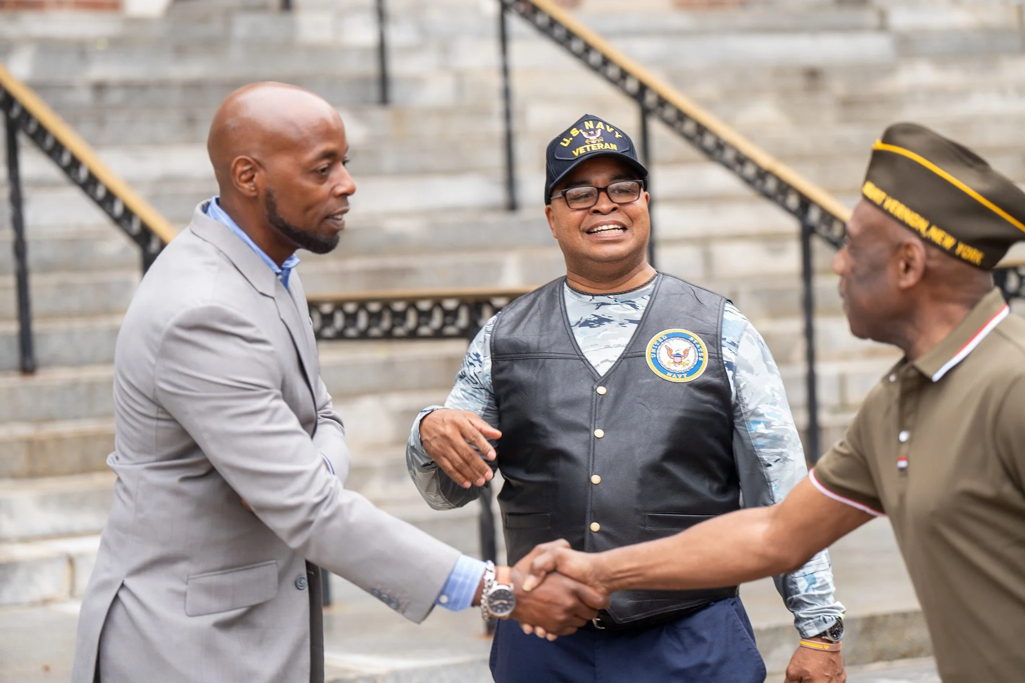 Three men, including one in a U.S. Navy vest and hat, shake hands and converse on a staircase outside.
