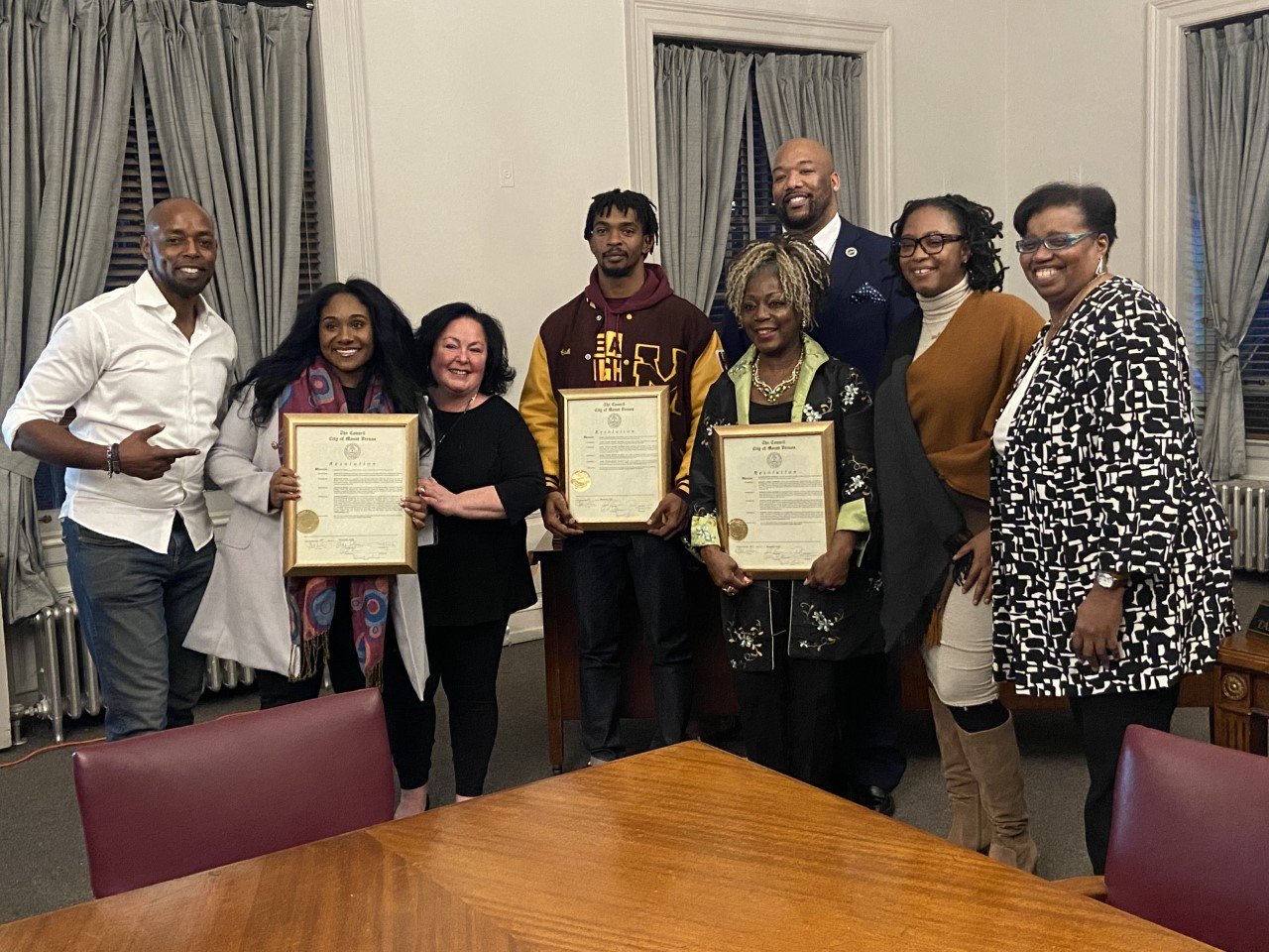 Group of nine people standing together in a room, some holding framed certificates, celebrating an achievement or award ceremony.