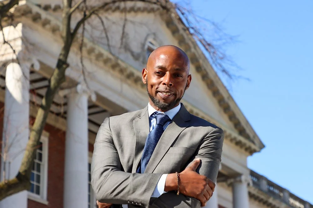 A man in a gray suit with a blue tie stands outside in front of a historic building with large columns, arms crossed, smiling at the camera.